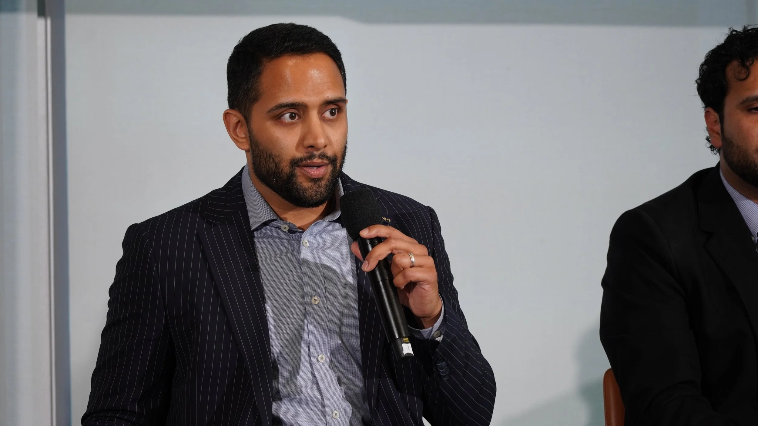 A man with a beard and dark hair, wearing a striped suit and a light gray shirt, holding a microphone and speaking during a panel discussion in an indoor conference setting.