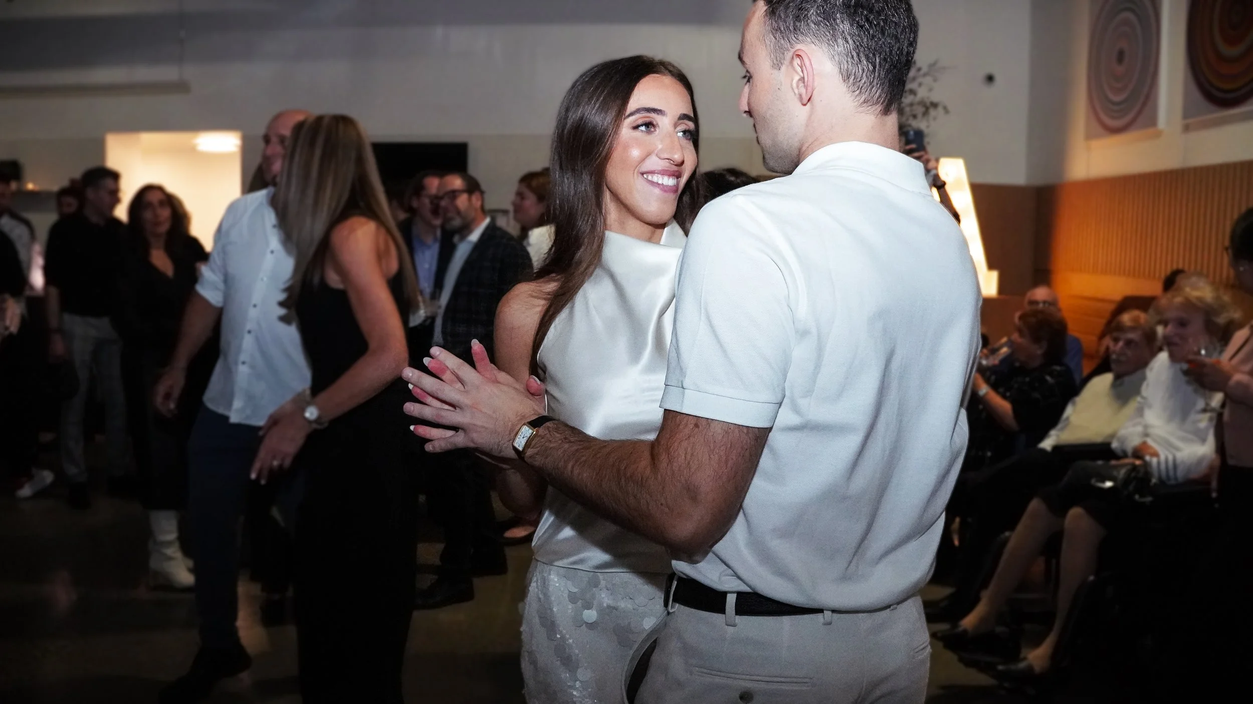 A woman and a man dance closely at a social event, smiling at each other. The woman is wearing a white dress, and the man is in a white shirt. People are gathered in the background, some sitting and some dancing.