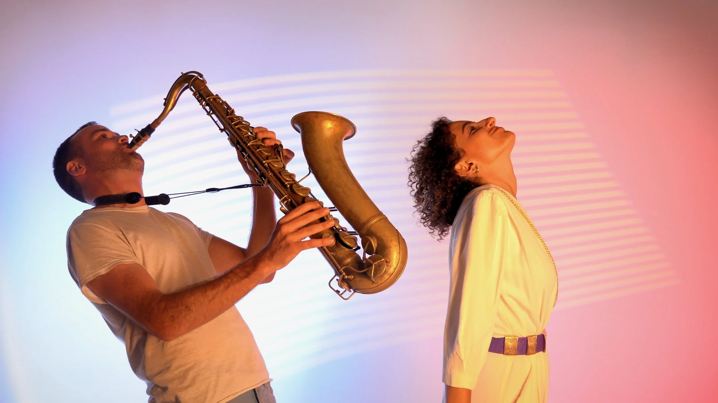 A man playing a saxophone with a woman standing nearby, both with their heads tilted back and eyes closed, under colorful, pink and blue lighting.