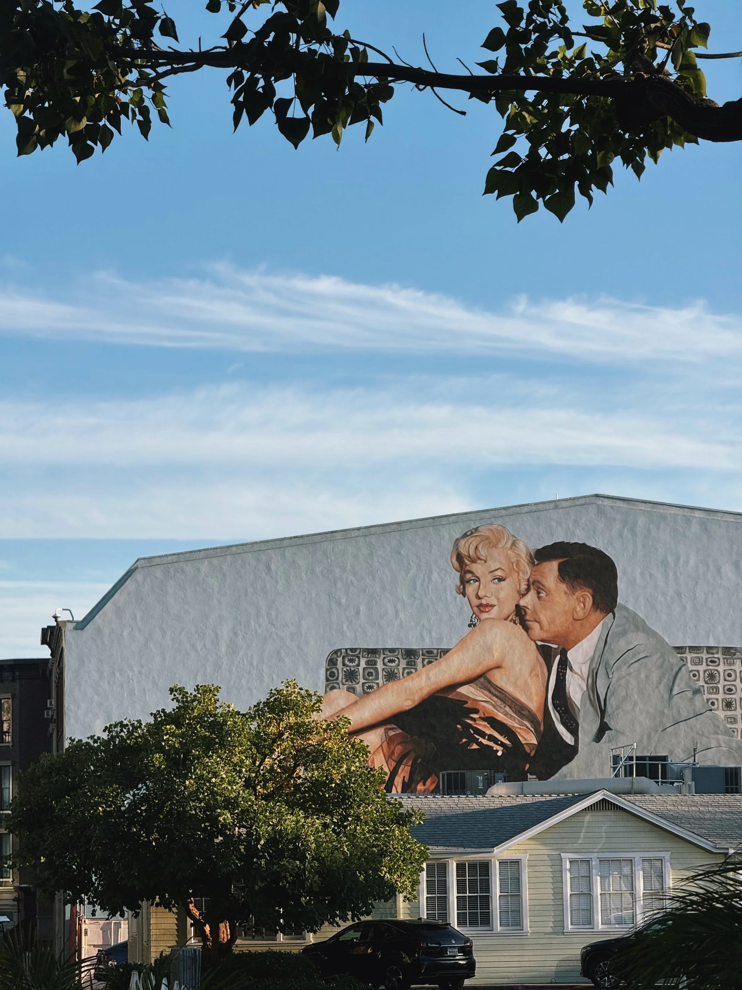 A mural on the side of a building depicting Marilyn Monroe and a man sitting closely together, with trees and houses in the foreground, and a blue sky with wispy clouds above.