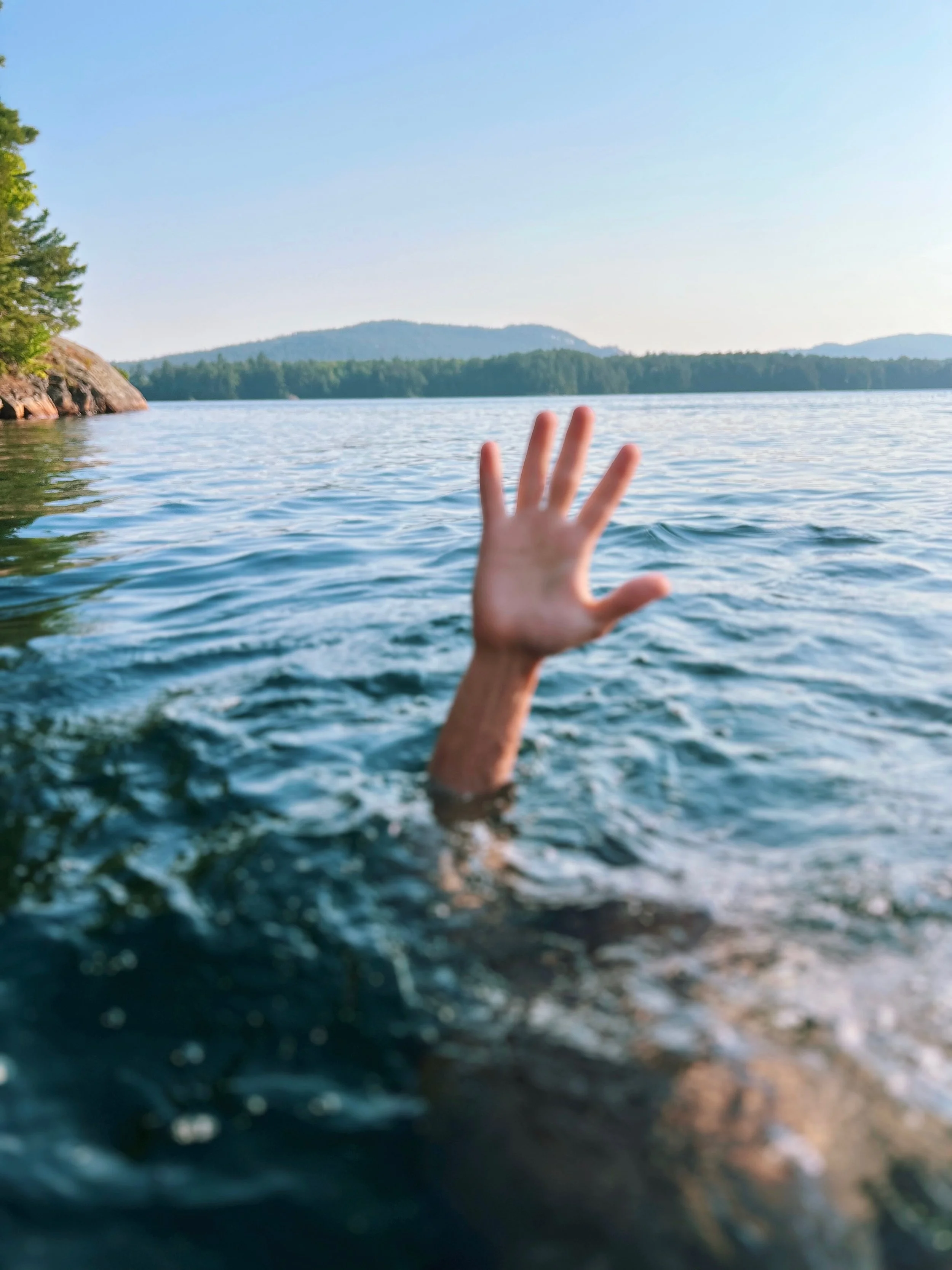 A person with an outstretched arm reaching above the water in a lake with a scenic backdrop of trees, hills, and a clear sky.