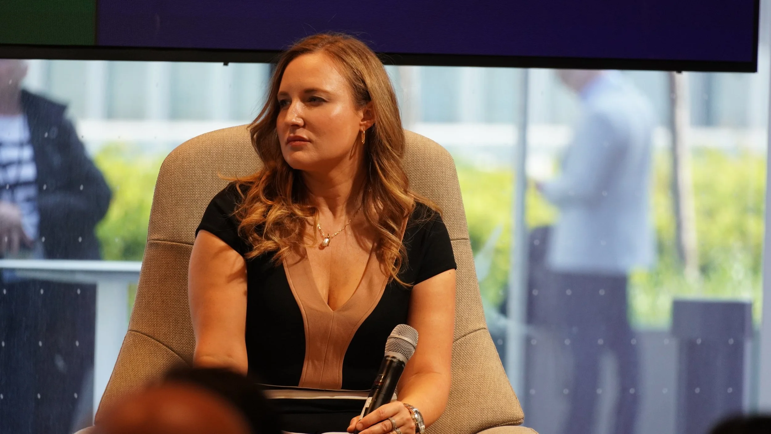 A woman with wavy red hair sitting in a beige chair, holding a microphone, in an indoor setting with large windows and blurred figures outside.