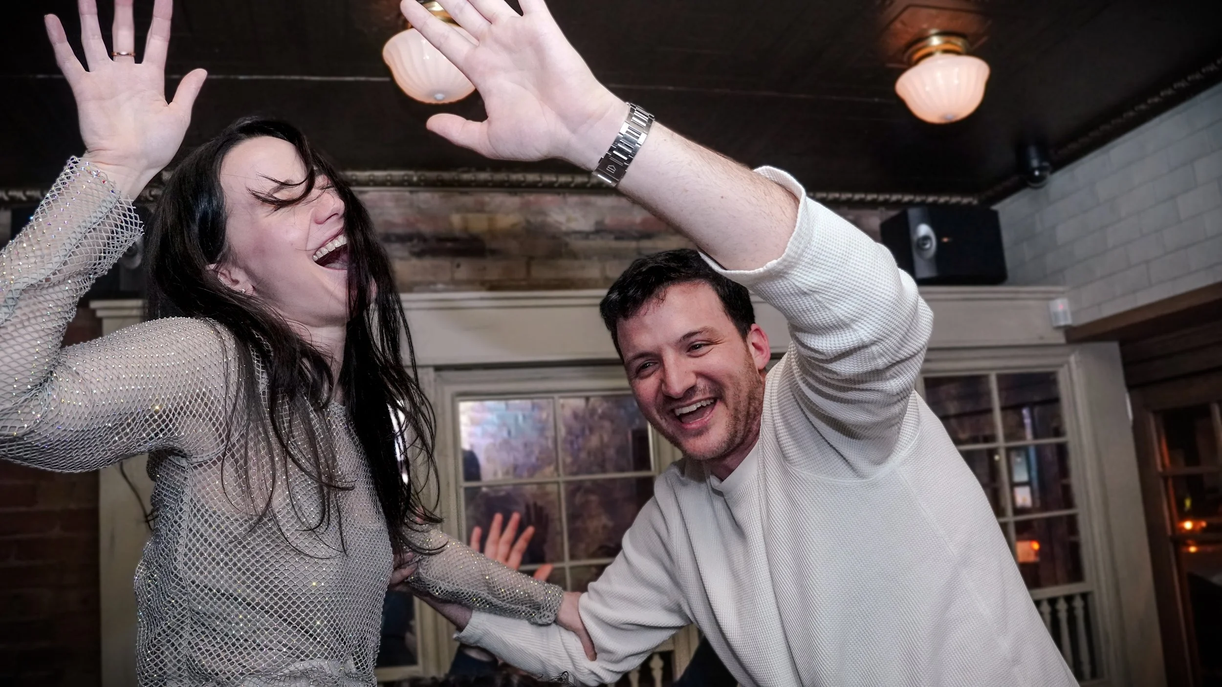 Two people dancing and smiling at a party, one woman with dark hair and a sparkly shirt, and one man with short dark hair and a white shirt, indoors with warm lighting.