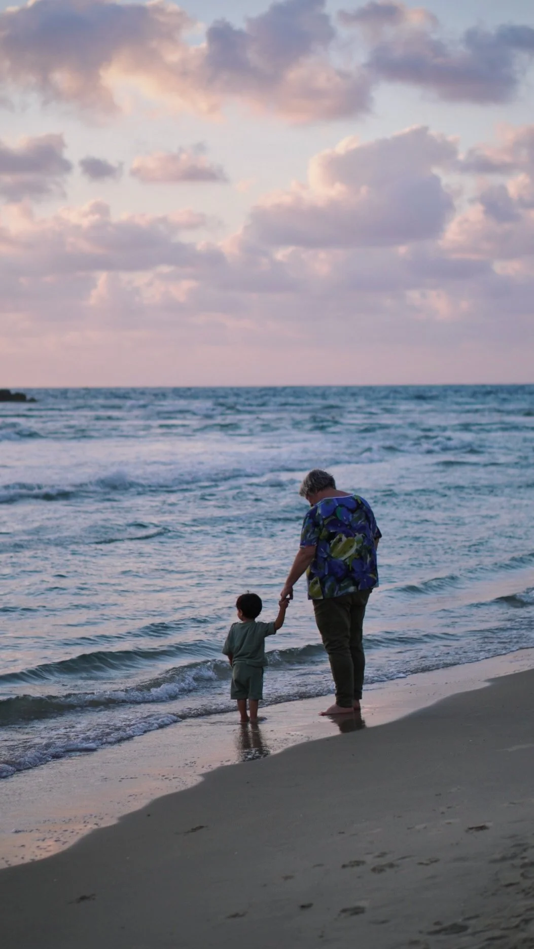 An adult and a child holding hands and walking along the beach at sunset, with ocean waves and clouds in the sky.