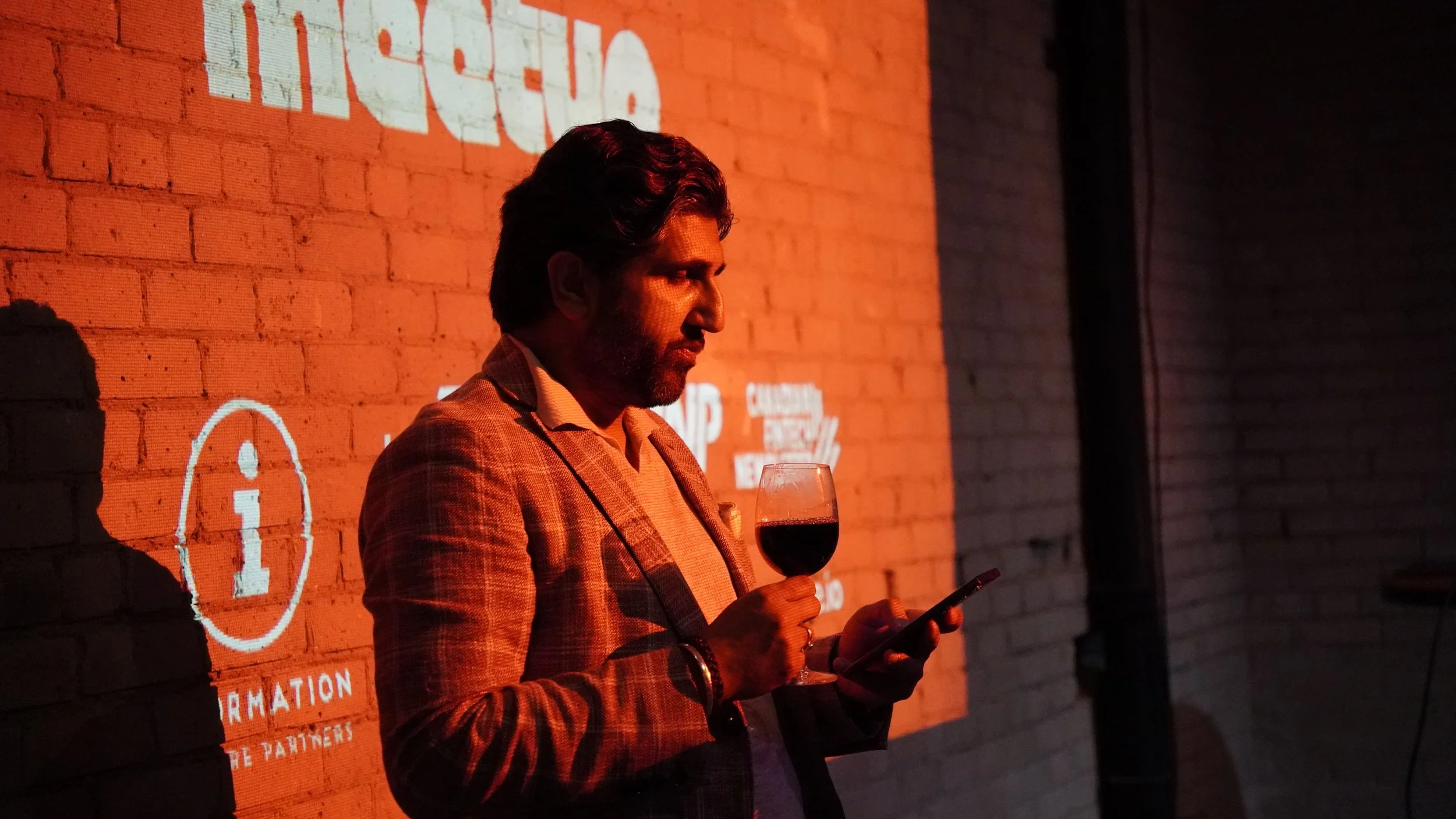 A man in a checkered blazer and light shirt is holding a glass of red wine in one hand and a smartphone in the other, standing against a brick wall with projected text and logos, dimly lit with warm tones.