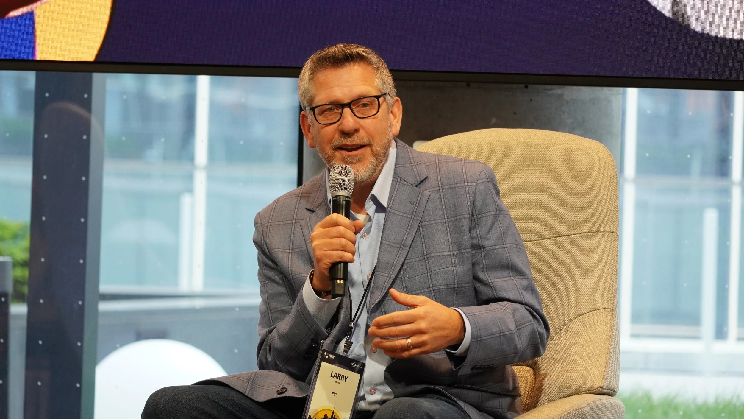 A middle-aged man wearing glasses and a gray plaid blazer sitting in a beige armchair with a microphone in his right hand, speaking at an indoor event.