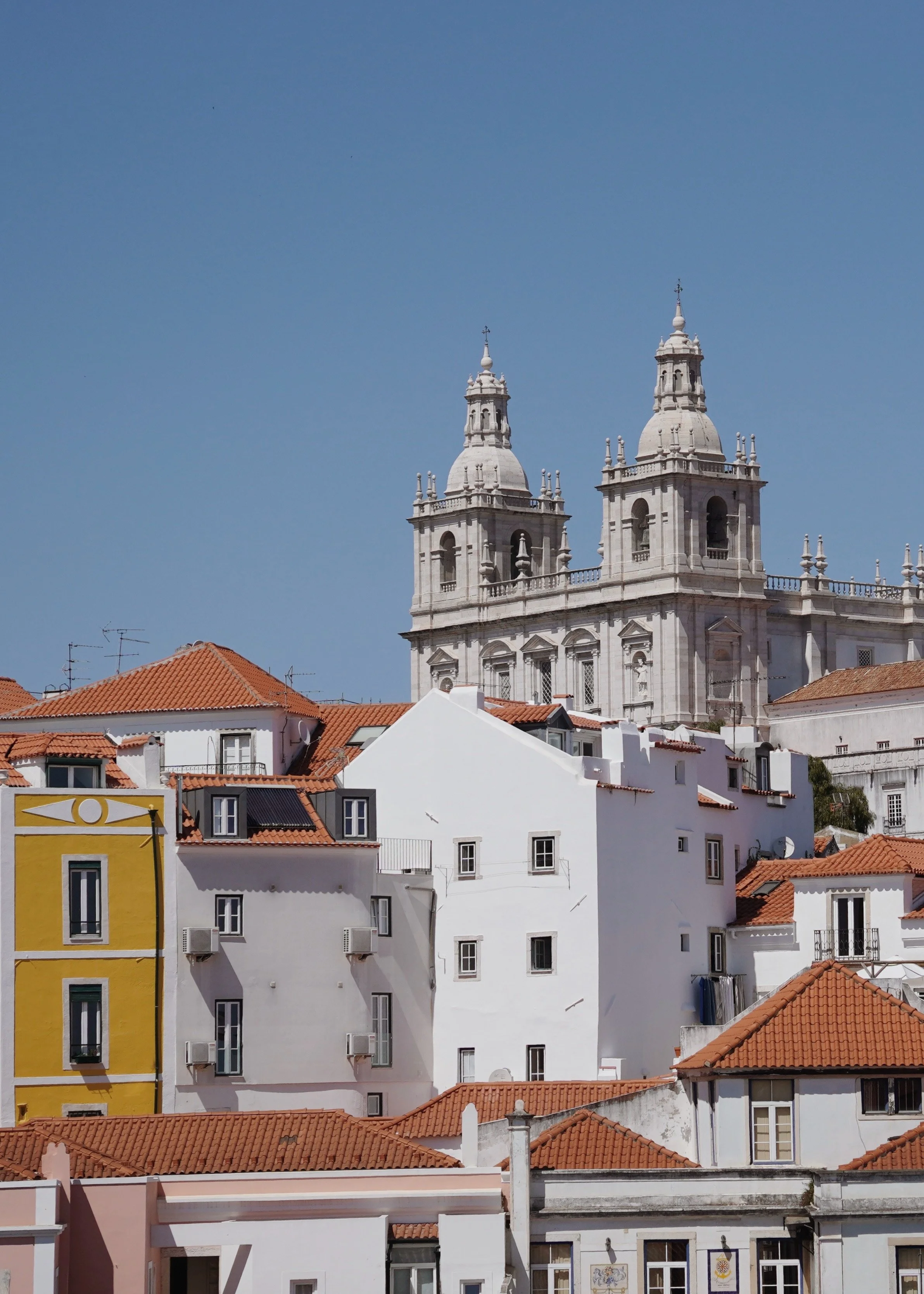 Cityscape with white buildings and red-tiled roofs in front of a large historical church with twin bell towers under a clear blue sky.