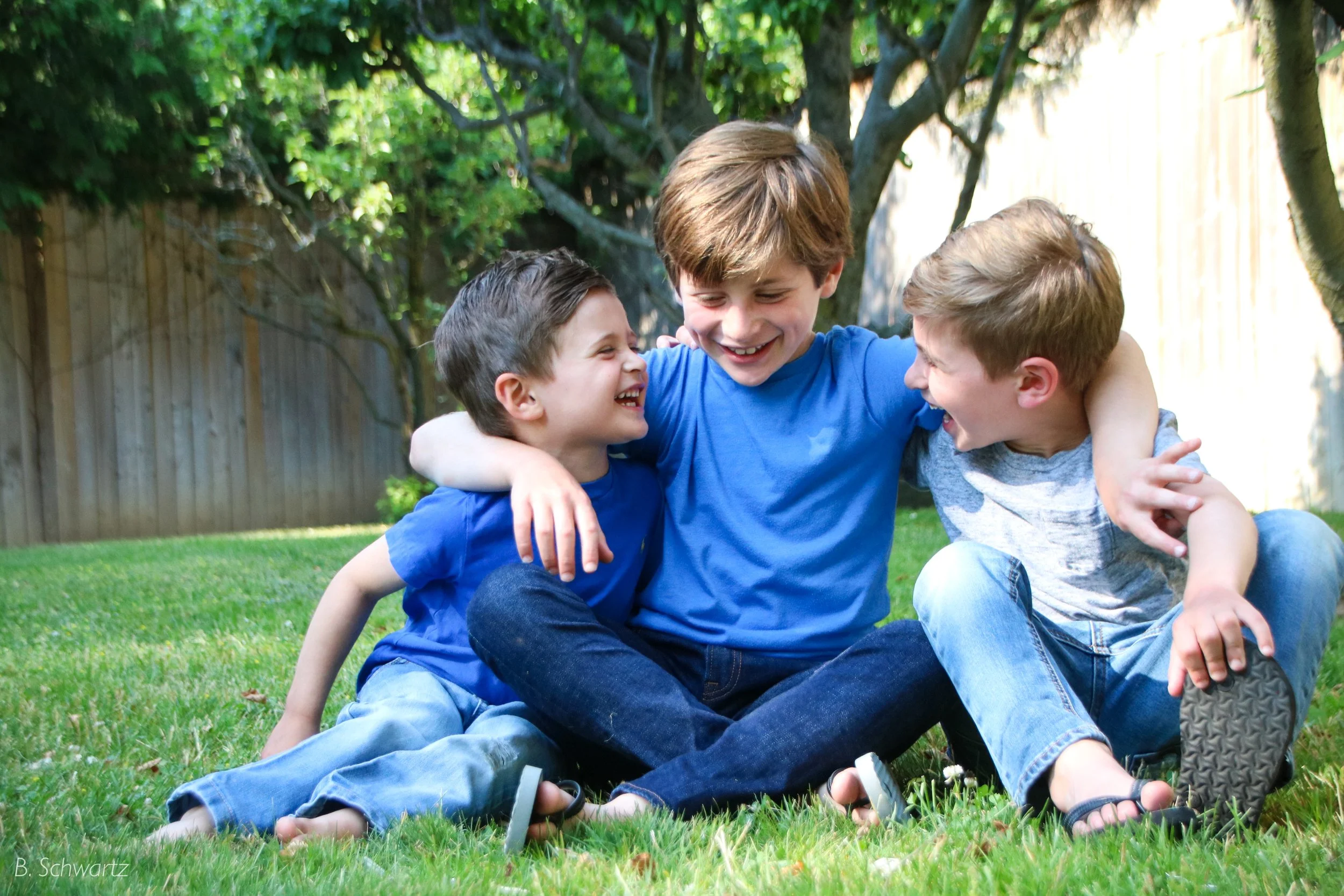 Three boys sitting on the grass in a backyard, laughing and hugging each other.
