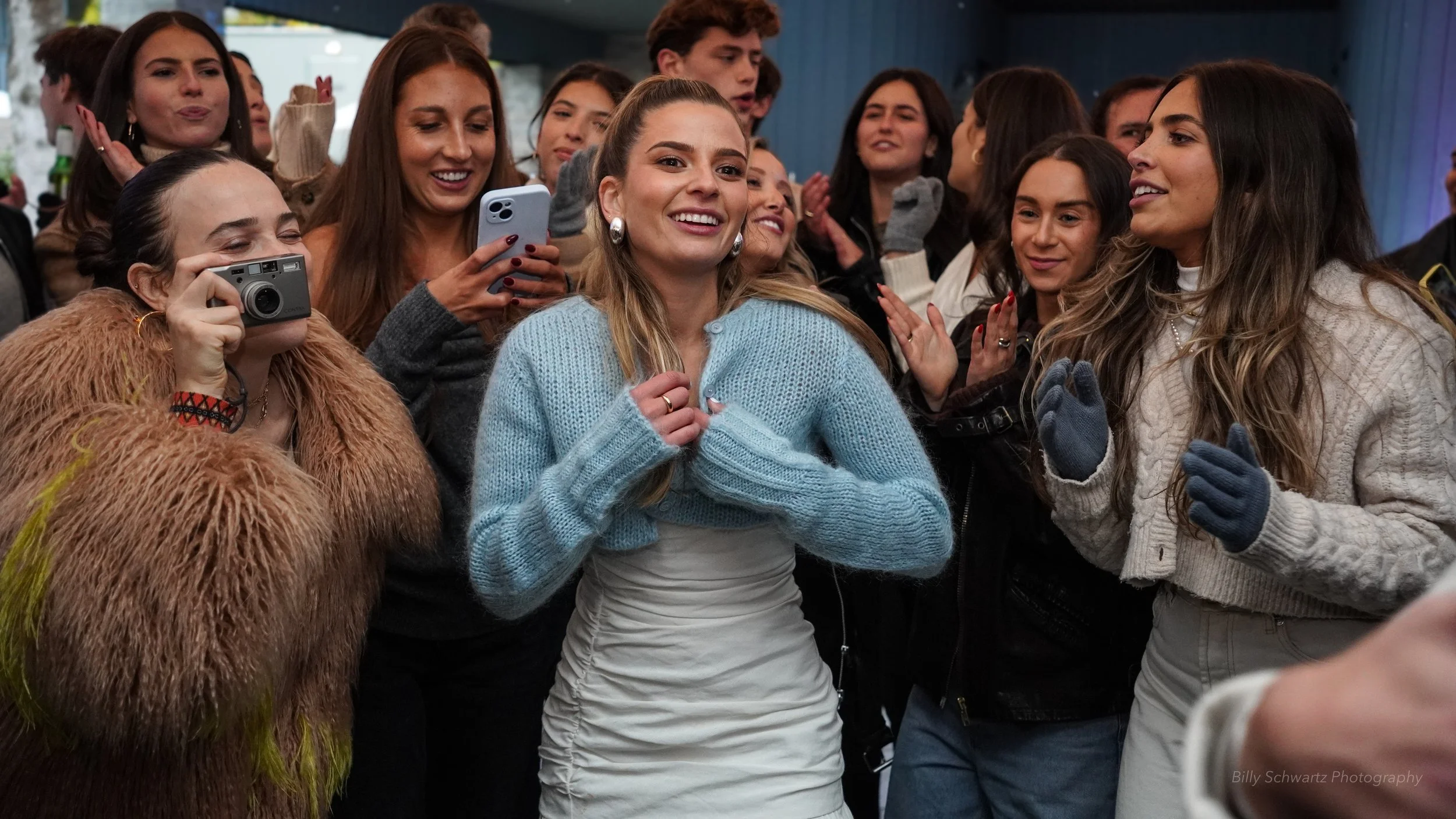 A group of women and girls are gathered closely together, smiling and taking photos. The woman in the center is wearing a light blue sweater and a white skirt, smiling with her hand on her chest. The woman on the right is wearing a beige knit sweater