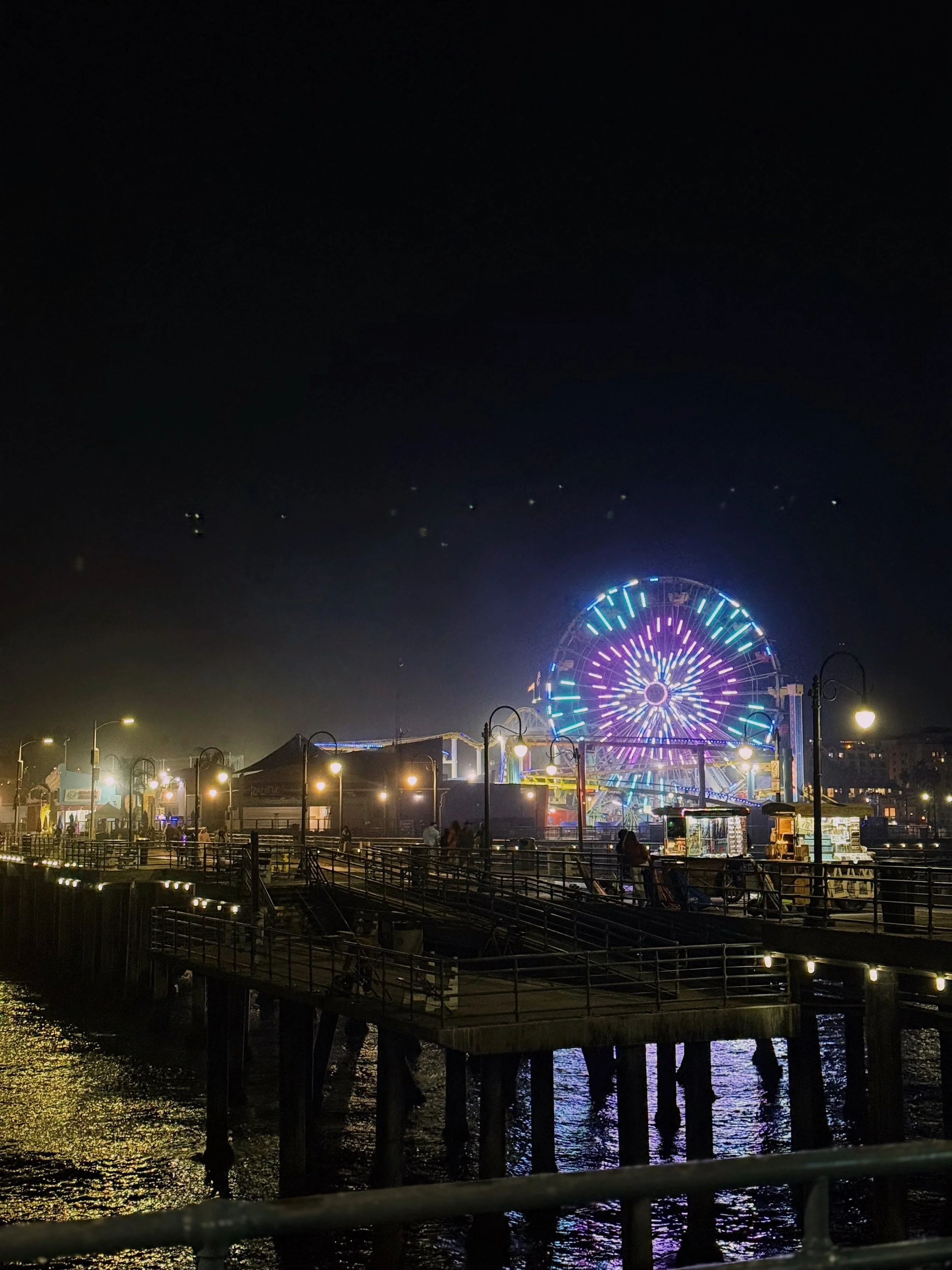 Nighttime view of a pier with a brightly lit Ferris wheel in the background at a seaside amusement park.