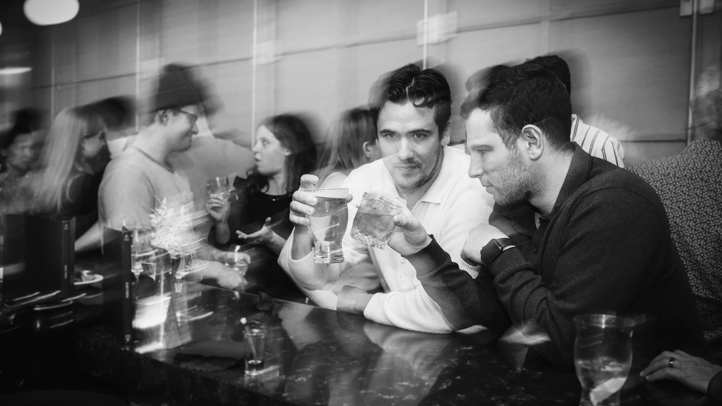 Two men sitting at a bar, one holding a glass of water and the other holding a drink in a glass. The scene is in black and white, with other people in the background.