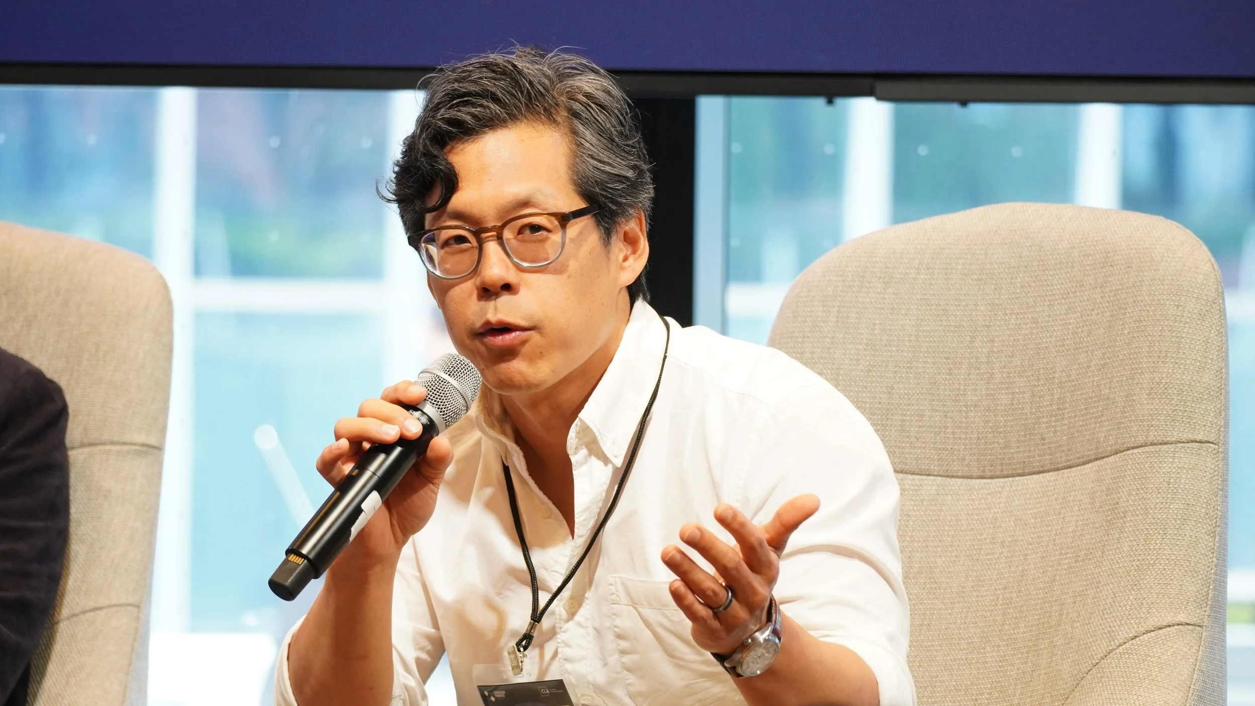 A man with black hair, glasses, wearing a white shirt, holding a microphone, speaking at a panel discussion or conference.