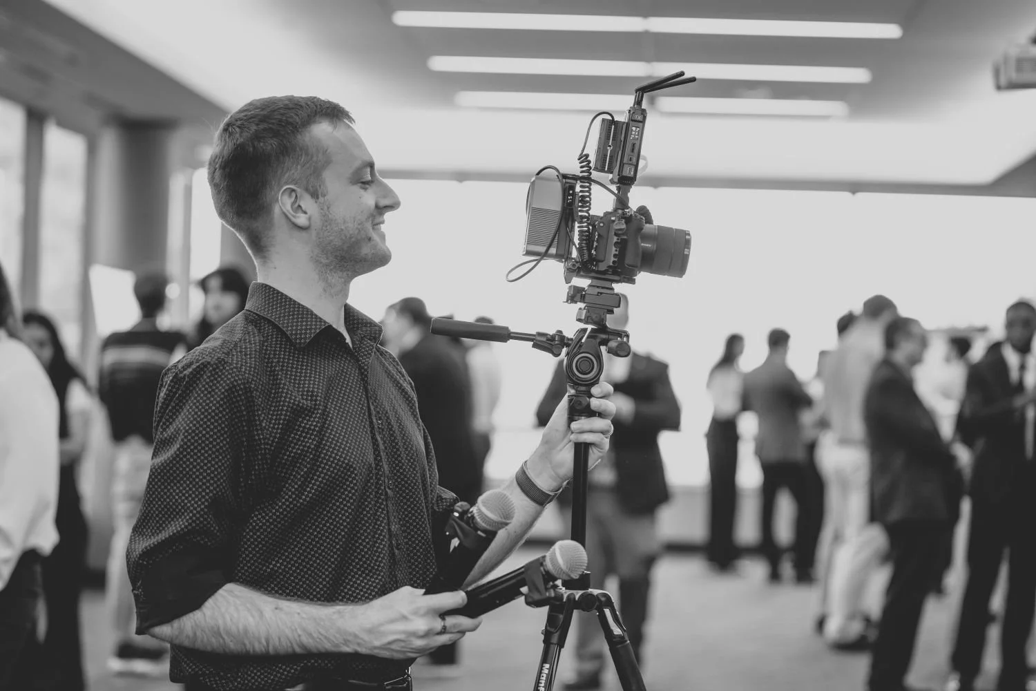 A man operating a professional camera mounted on a stabilizer, capturing footage at an indoor event with several people in the background.