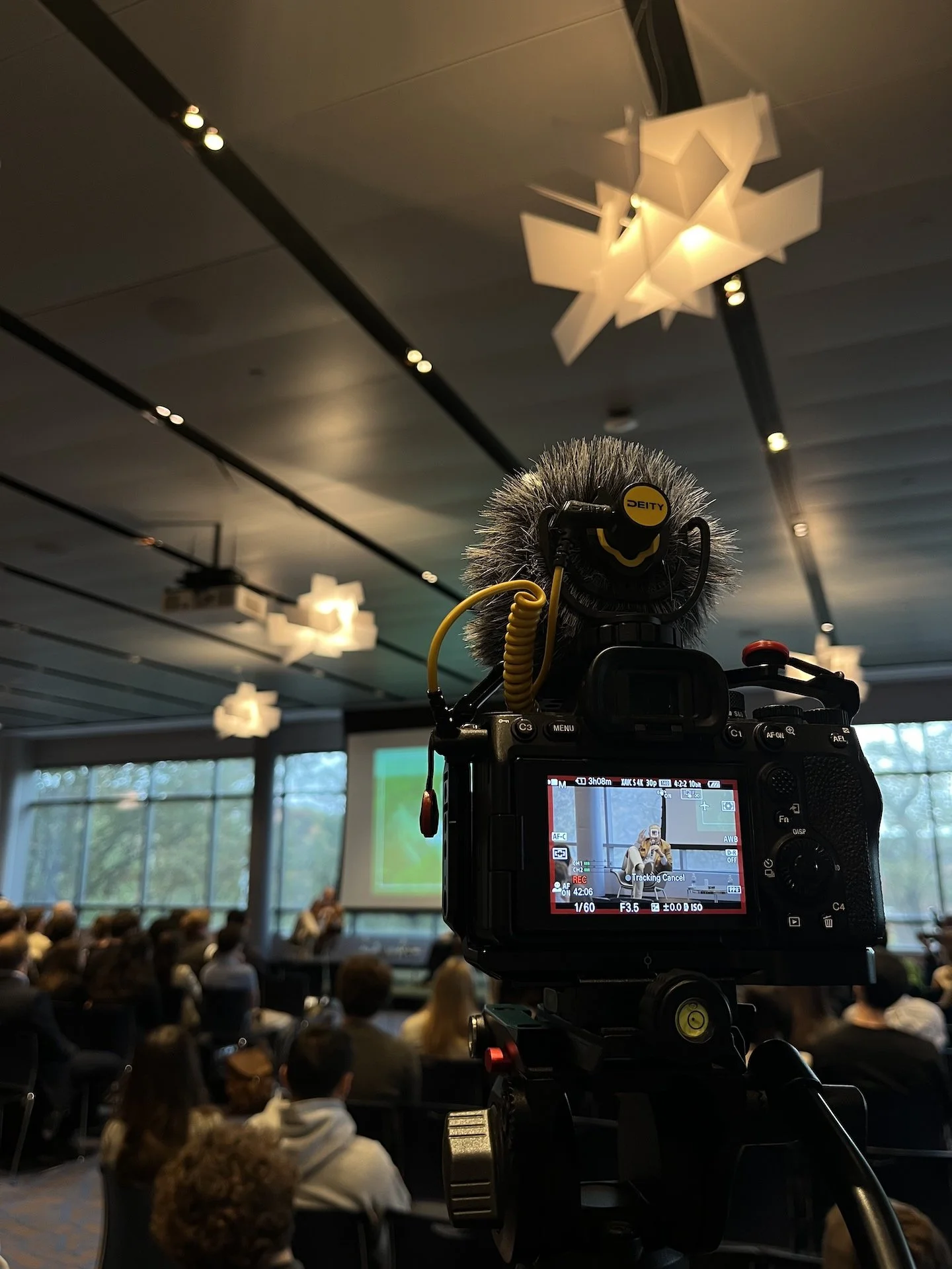 A digital camera on a tripod capturing a presentation at a conference room filled with seated attendees. The room has large windows and ceiling lights, with a large screen displaying slides at the front.