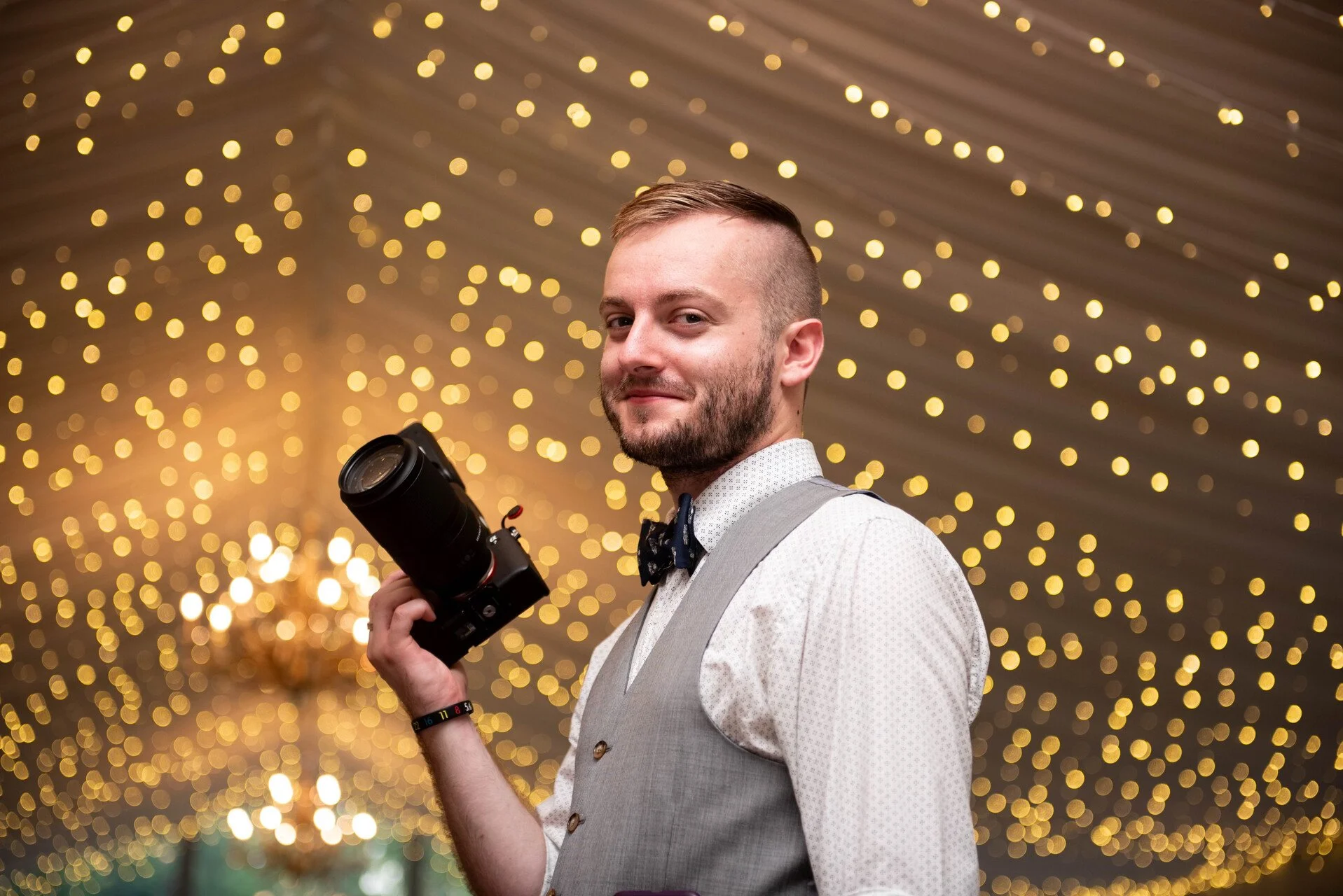 A man dressed in a light-colored vest and dress shirt, holding a camera, with a background of warm, yellow string lights creating a bokeh effect. He has short hair and a beard, and is looking at the camera with a slight smile.