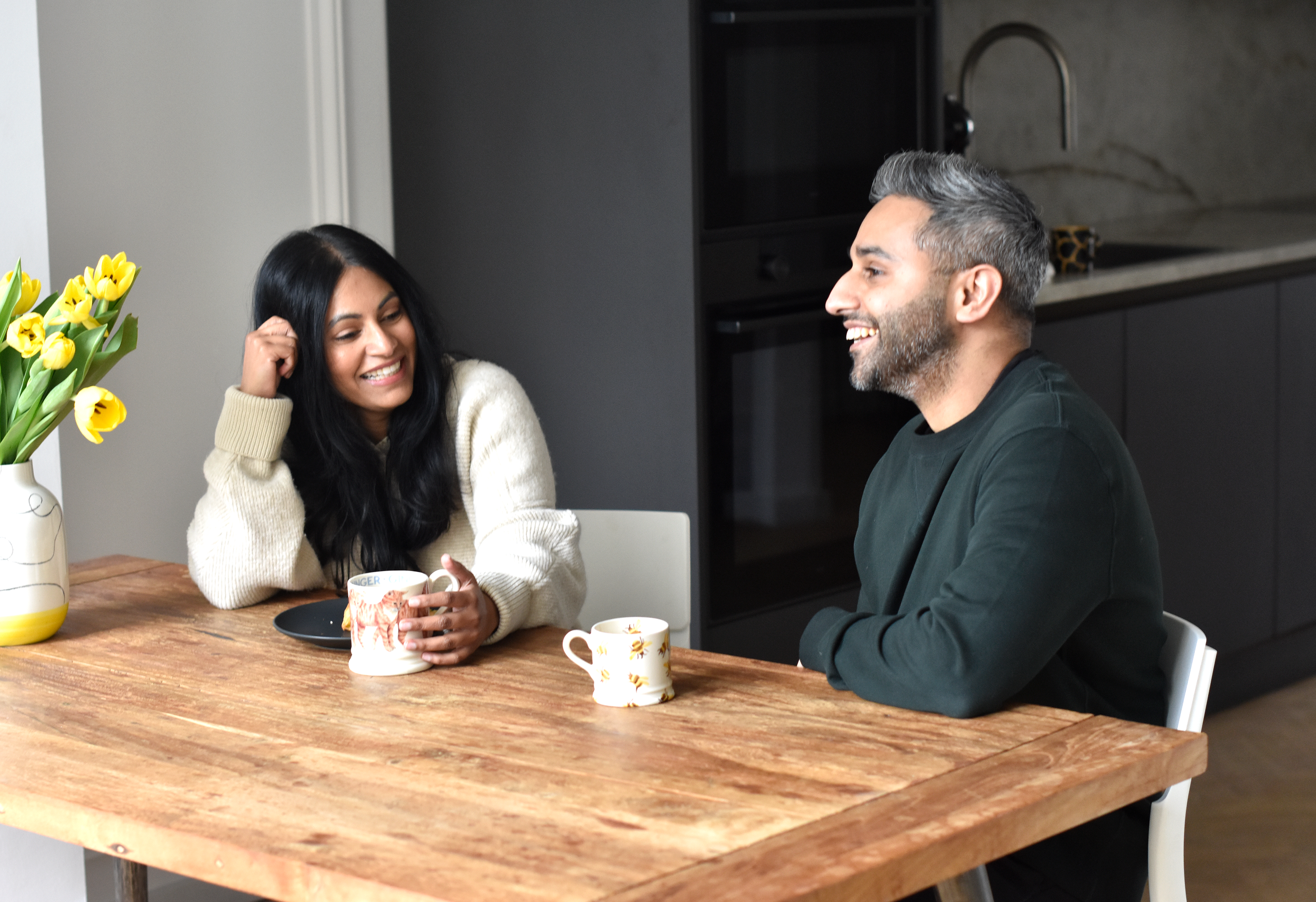 A woman and a man sitting at a wooden table, smiling and having a conversation. The woman has long black hair and is wearing a cream sweater, holding a mug. The man has gray hair and a beard, wearing a dark shirt. There are two mugs and a vase with yellow tulips on the table, with a modern kitchen in the background.