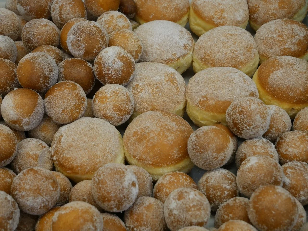 Nahaufnahme einer Bäcker-Auslage mit vielen kleinen, mit Puderzucker bestäubten Mini-Quarkbällchen links und größeren, ebenfalls puderzuckerbestäubten Berlinern rechts, dicht beieinander gestapelt.
