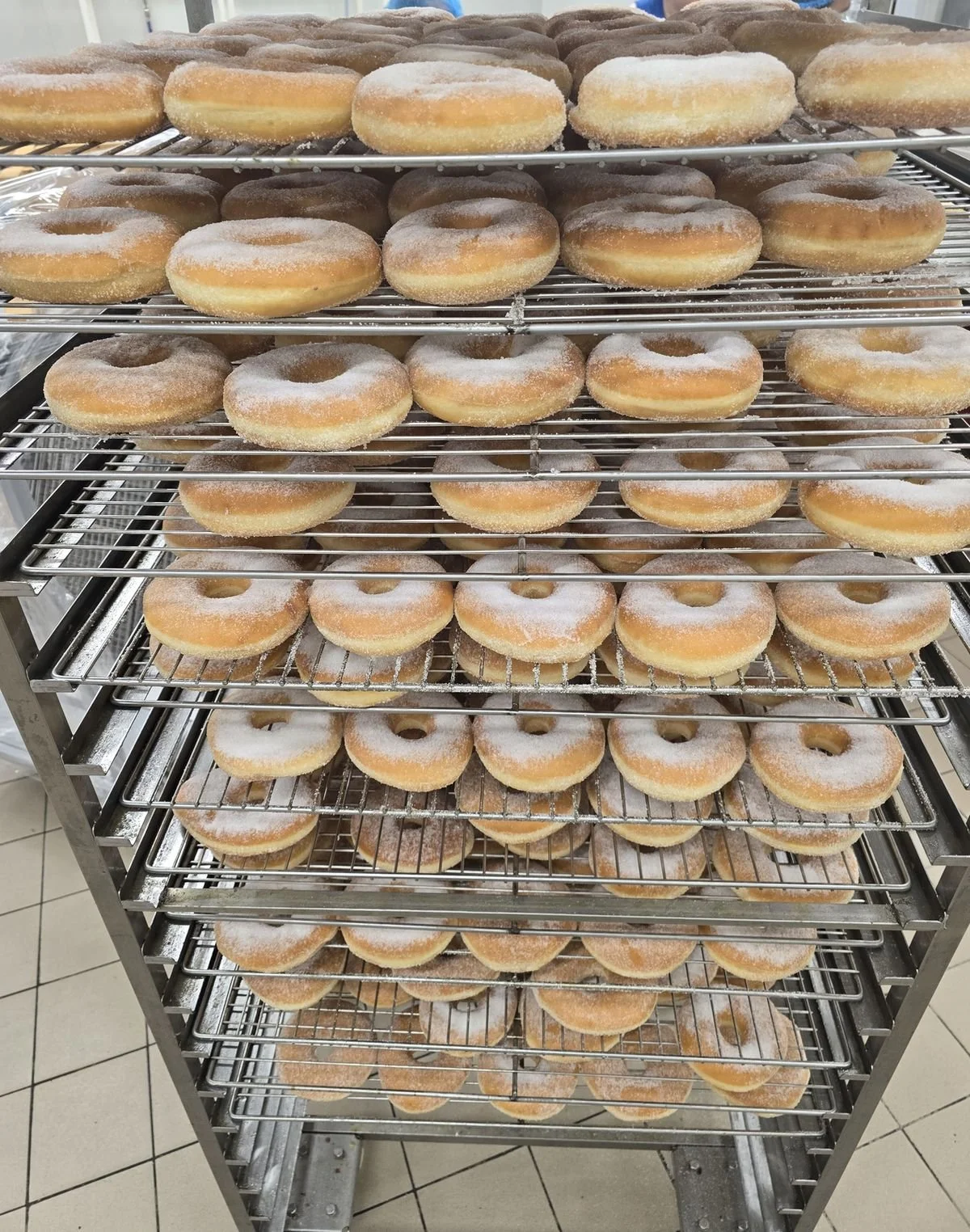Tall metal cooling rack filled with large sugar-dusted Greek Loukoumas pastries arranged on multiple shelves in a bakery.