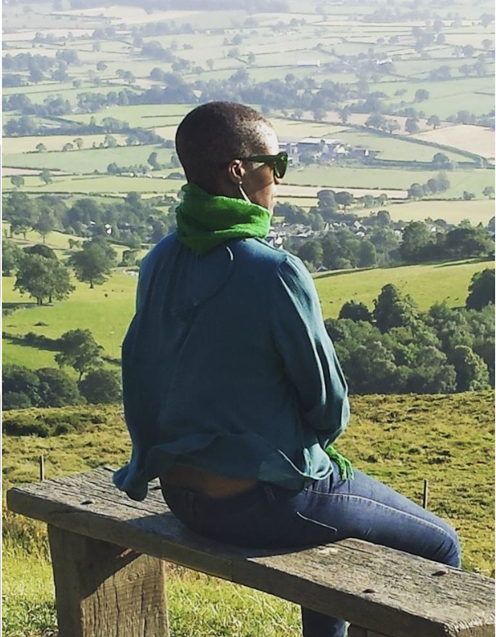 Jennifer John sitting on a wooden bench in a countryside landscape, back to the camera wearing sunglasses, a green scarf and earphones, facing beautiful lush Welsh countryside
