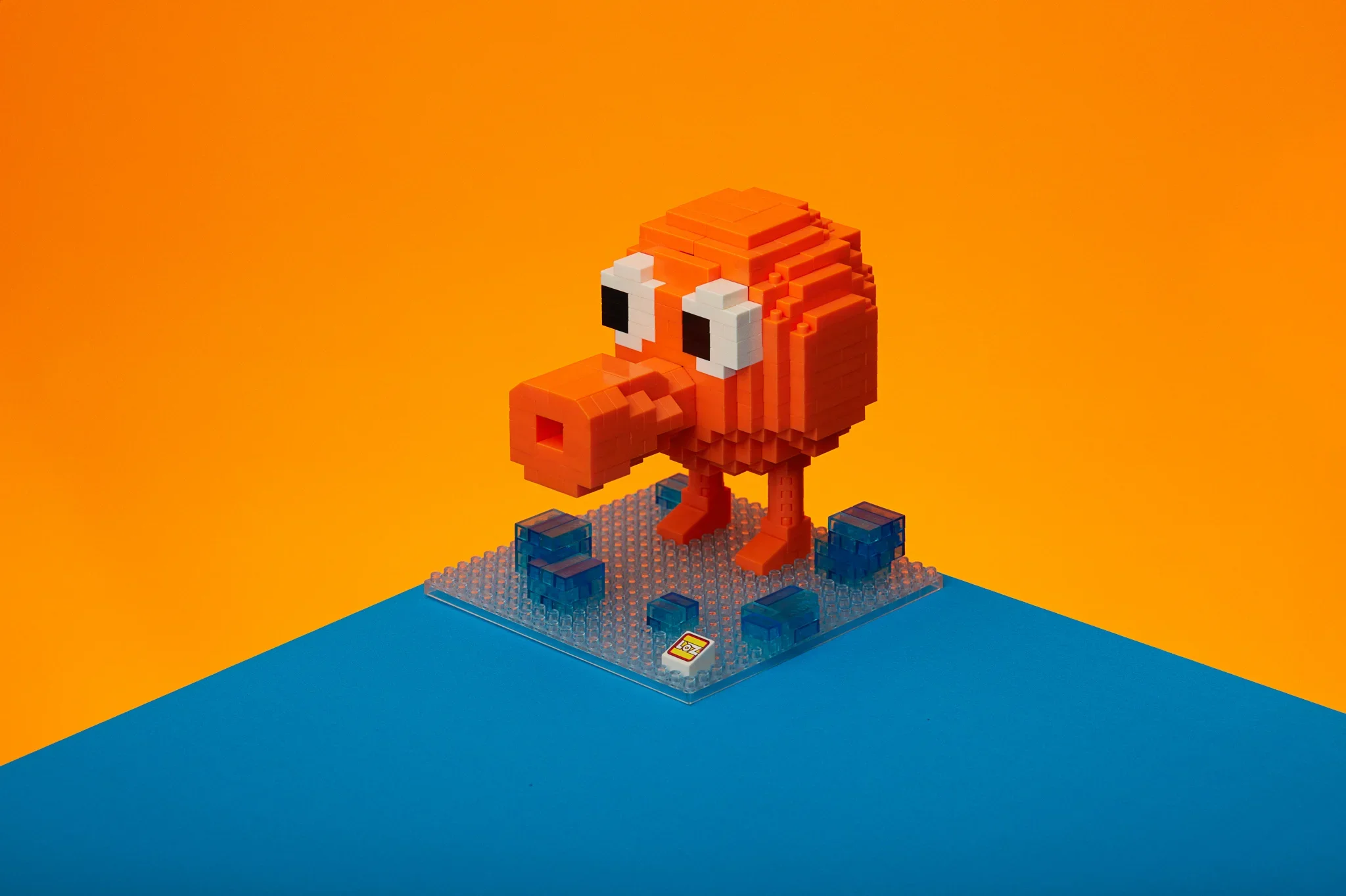 A pixel art sculpture of a red and white creature, resembling Bowser from the Mario video game series, made from building blocks, standing on a platform with blue and yellow backgrounds.