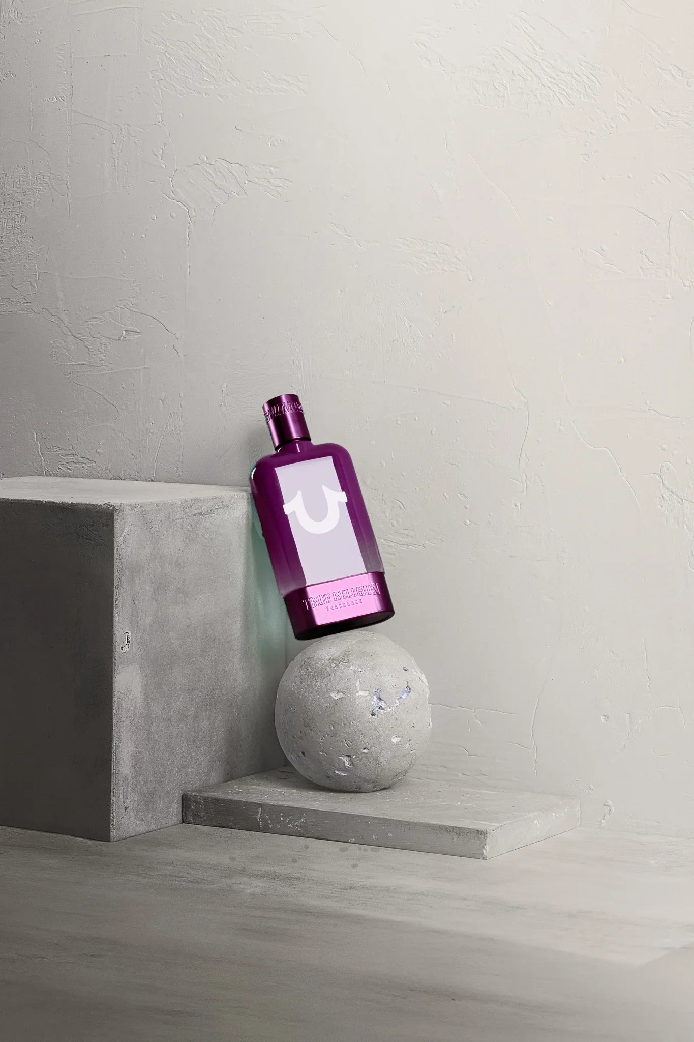 A purple bottle of True Religion fragrance resting on a spherical cement sculpture, set against a textured off-white wall background with concrete blocks.