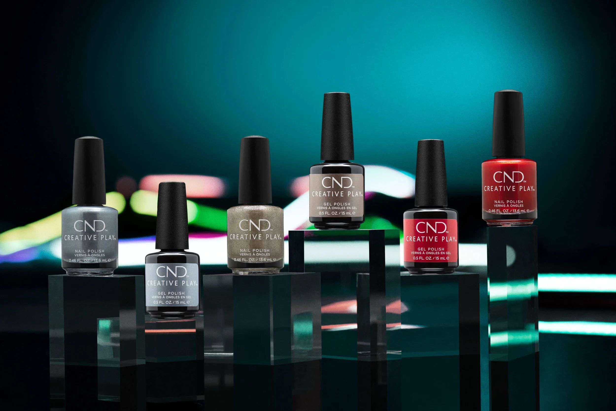 Six bottles of CND Creative Play gel nail polish displayed on black stands against a blurred colorful background. The bottles are in shades of gray, beige, and red with black caps.