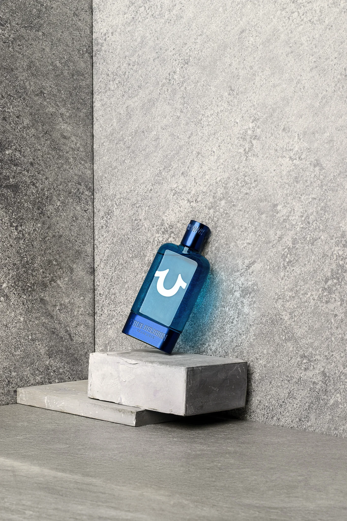 A blue bottle of True Religion Fragrance rests on a white concrete block in front of a textured, two-tone gray background. The bottle is placed diagonally.