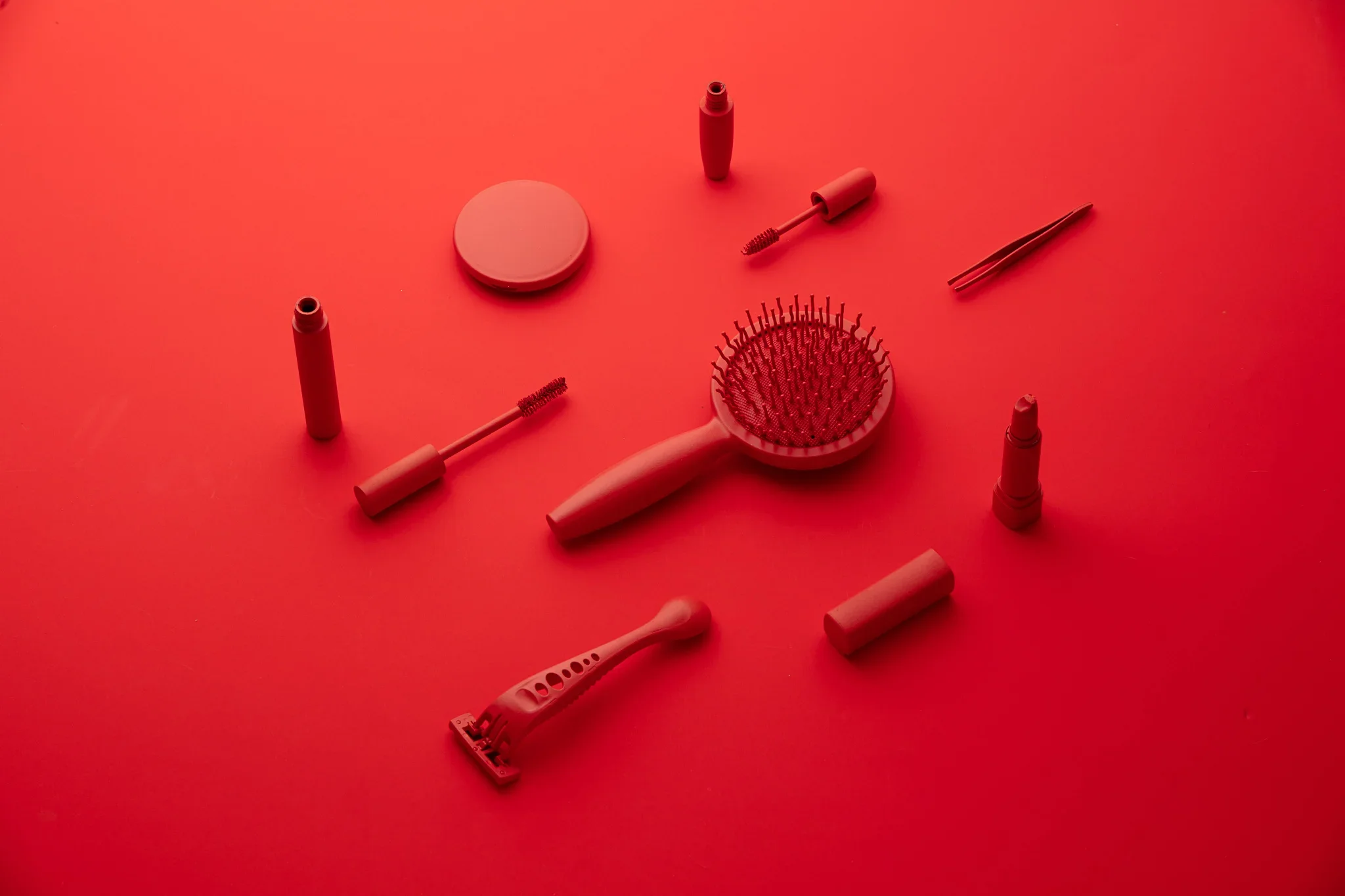 Various beauty and grooming tools including a round mirror, a mascara wand, a toilet brush, a pair of tweezers, a lipstick, an eyeshadow palette, an electric razor, and a nail clipper, all arranged on a red background.