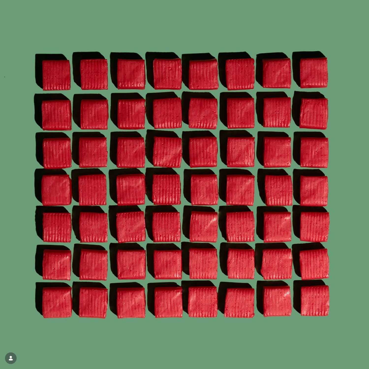 A grid of red square chocolates with a textured surface on a green background, casting shadows.