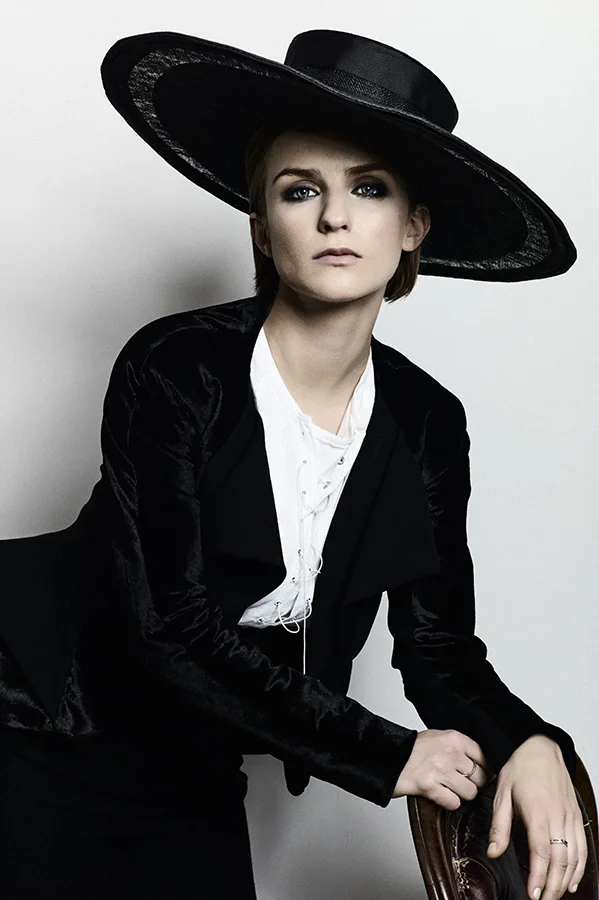 Faye wears whit tunic shirt with lace-up detailing by Shao Yen, ribbed black long waistcoad by James Lakeland, black high waist wide leg trousers by Rawan B, hat by Monique Lee Millinery
