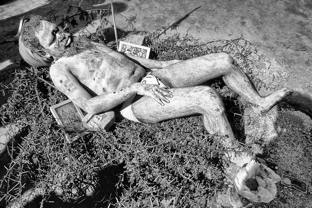 Black-and-white photograph of a Naga Sadhu reclining on a bed of thorn branches, with devotional pictures beside him, at the 1989 Maha Kumbh Mela in Allahabad, India.