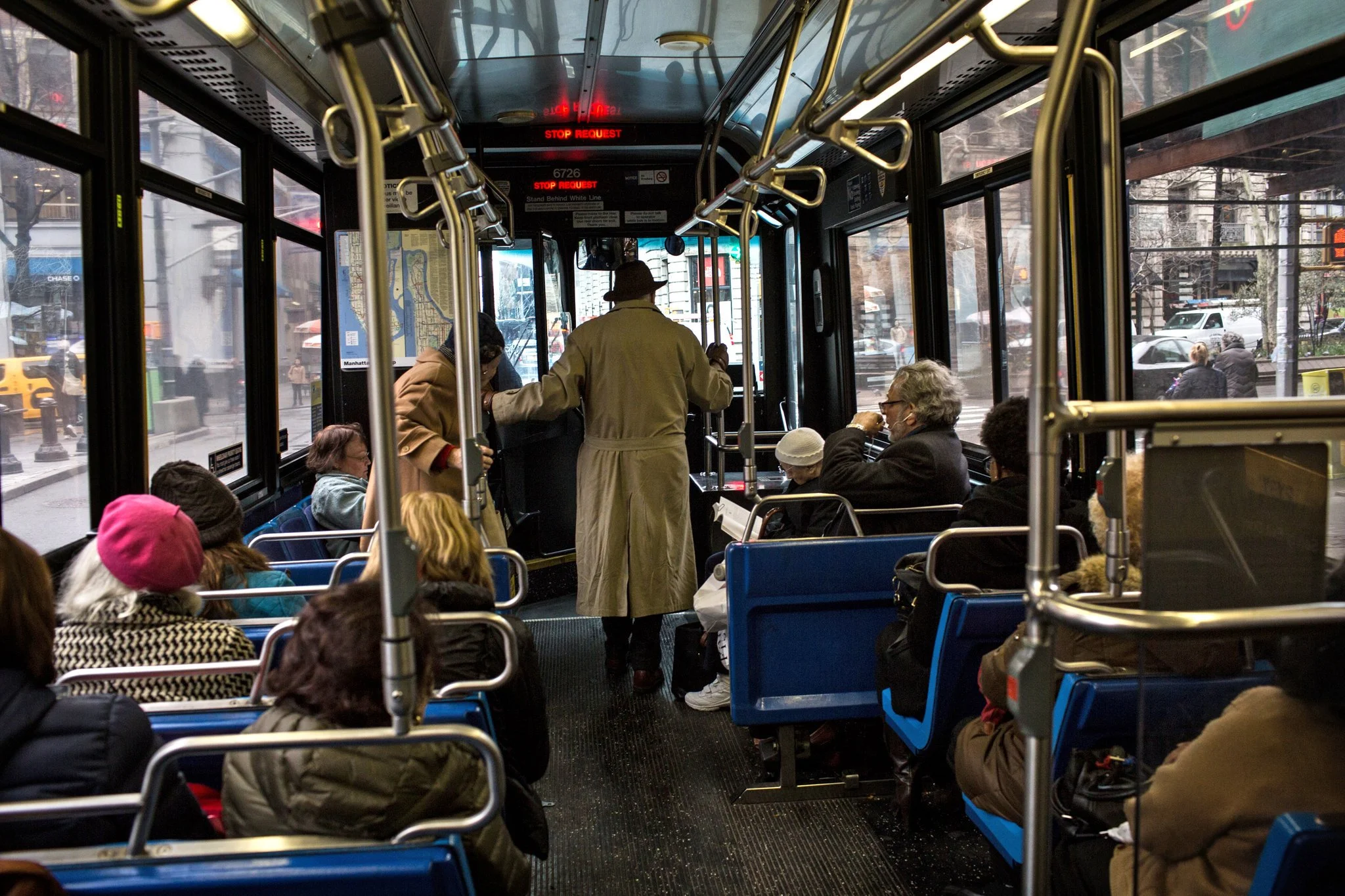 Inside a city bus with passengers seated and standing, some wearing winter clothing, and a driver at the front. Outside city streets and buildings are visible through the windows.