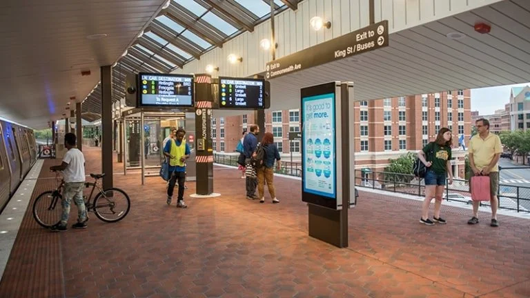 People walking and standing at a train station, with WMATA digital displays showing train schedules and information.