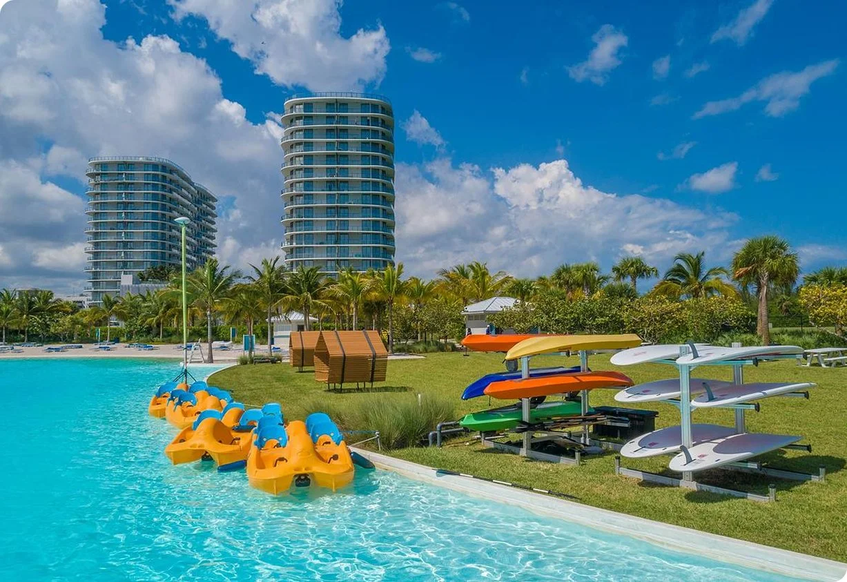 Resort pool with paddle boats, surfboards, and high-rise buildings under blue sky.
