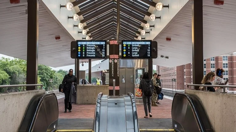People descend an escalator at a train station, with WMATA digital displays visible in the background.