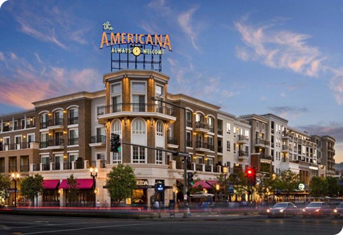 Exterior of The Americana at Brand shopping center during evening with shops and residential buildings under a twilight sky.