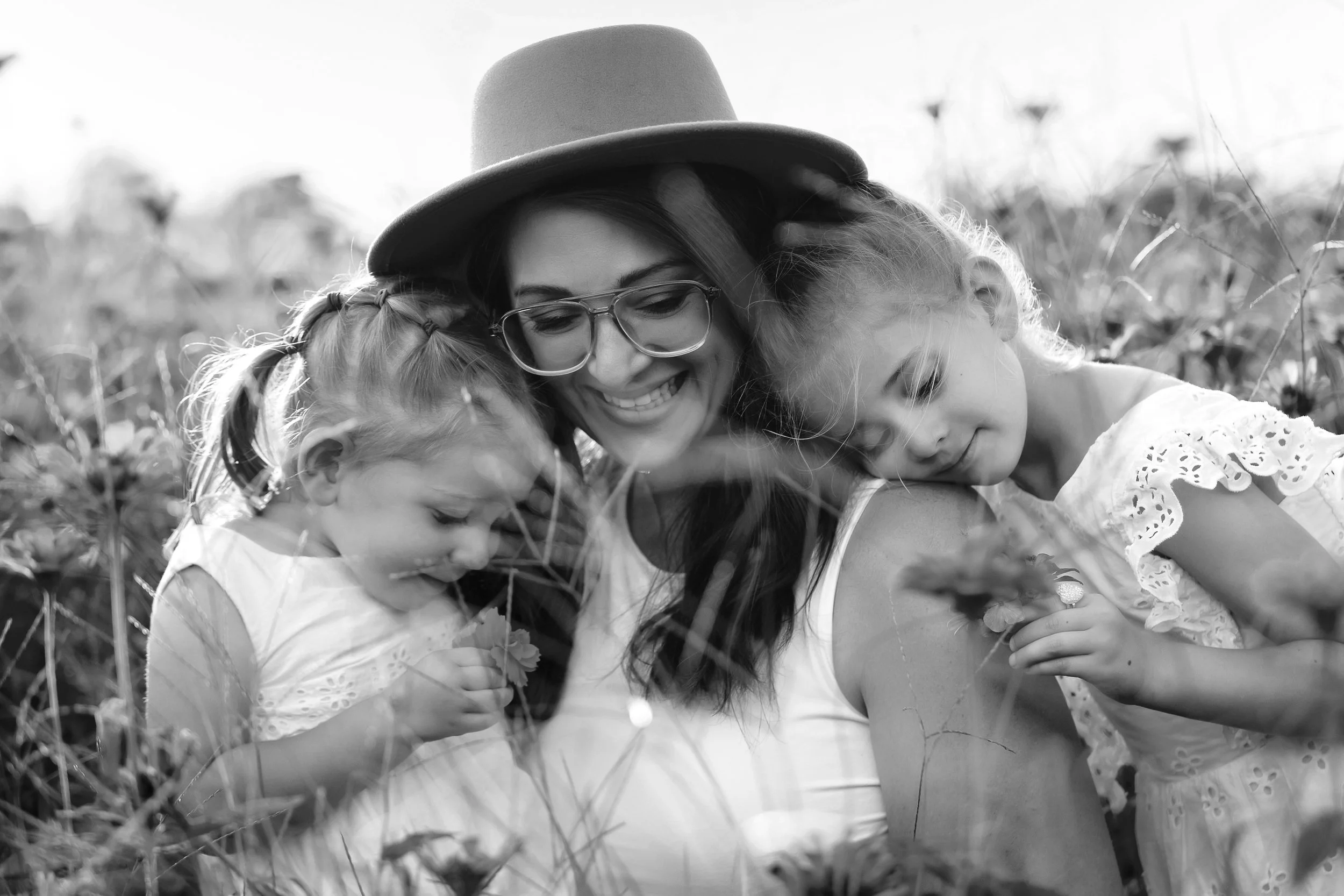 A woman with glasses and a hat smiling as she hugs two young girls, all three are in a field with tall grass and wildflowers, in black and white.