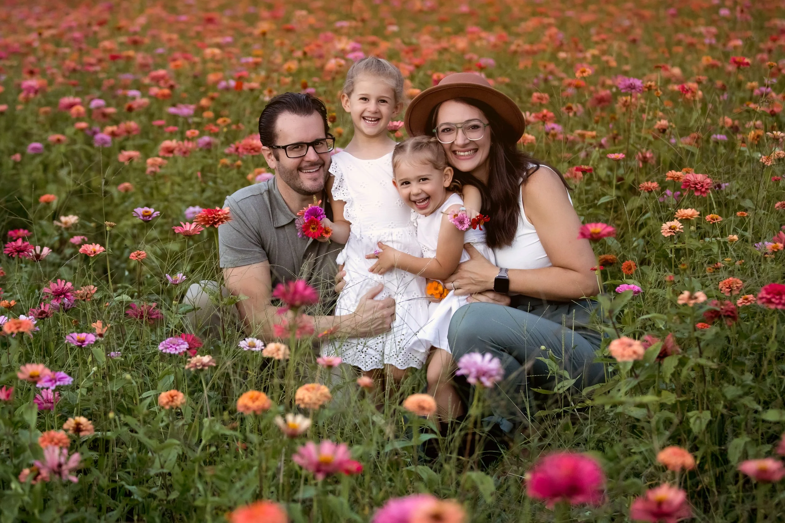 A happy family of five posing in a colorful flower field, with flowers in pink, purple, orange, and red shades.