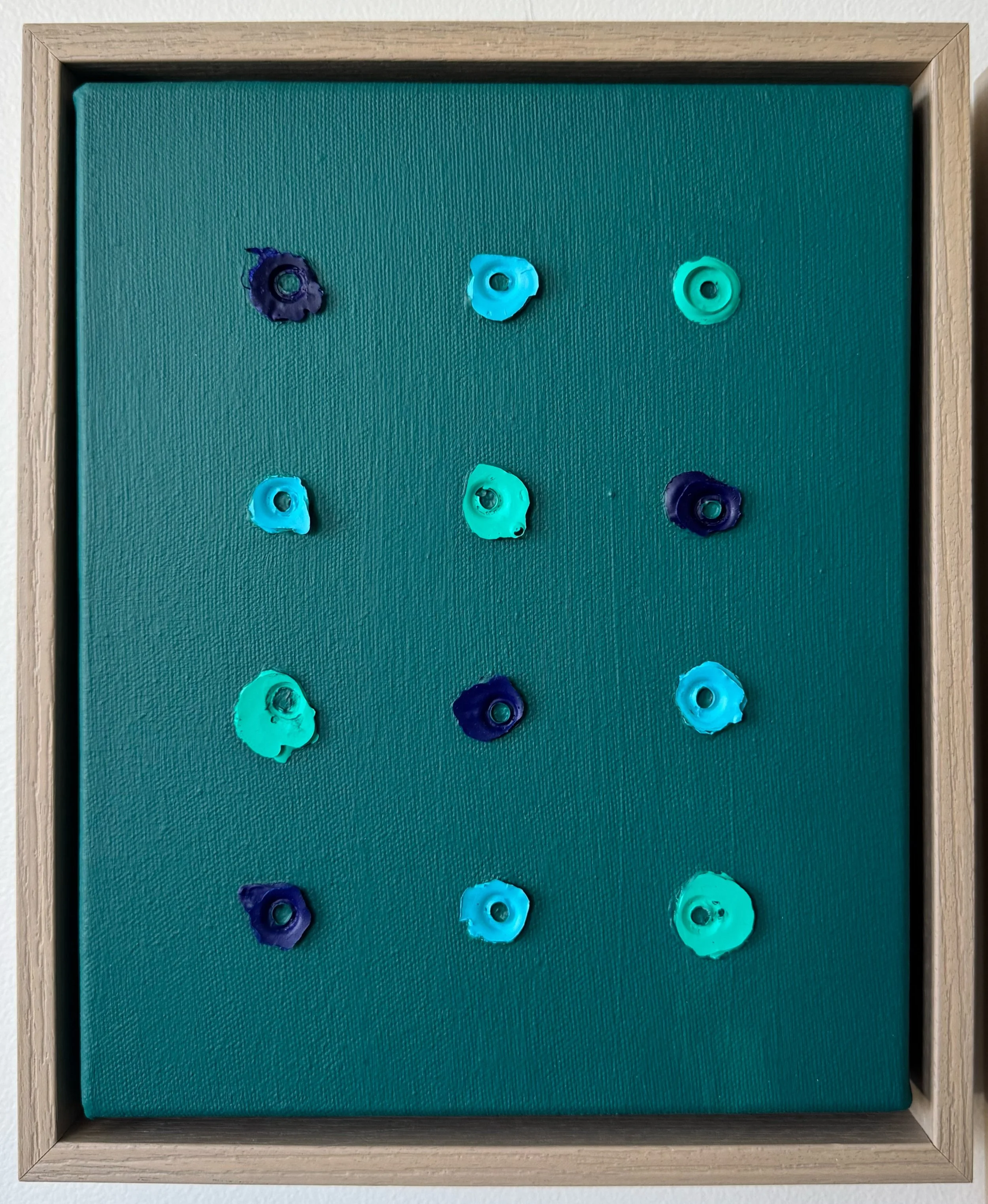 A textured teal painted background with twelve small, colorful, organic, circular dried pieces of acrylic paint arranged in a grid pattern, each with a hole in the center. The objects are in shades of blues and greens.