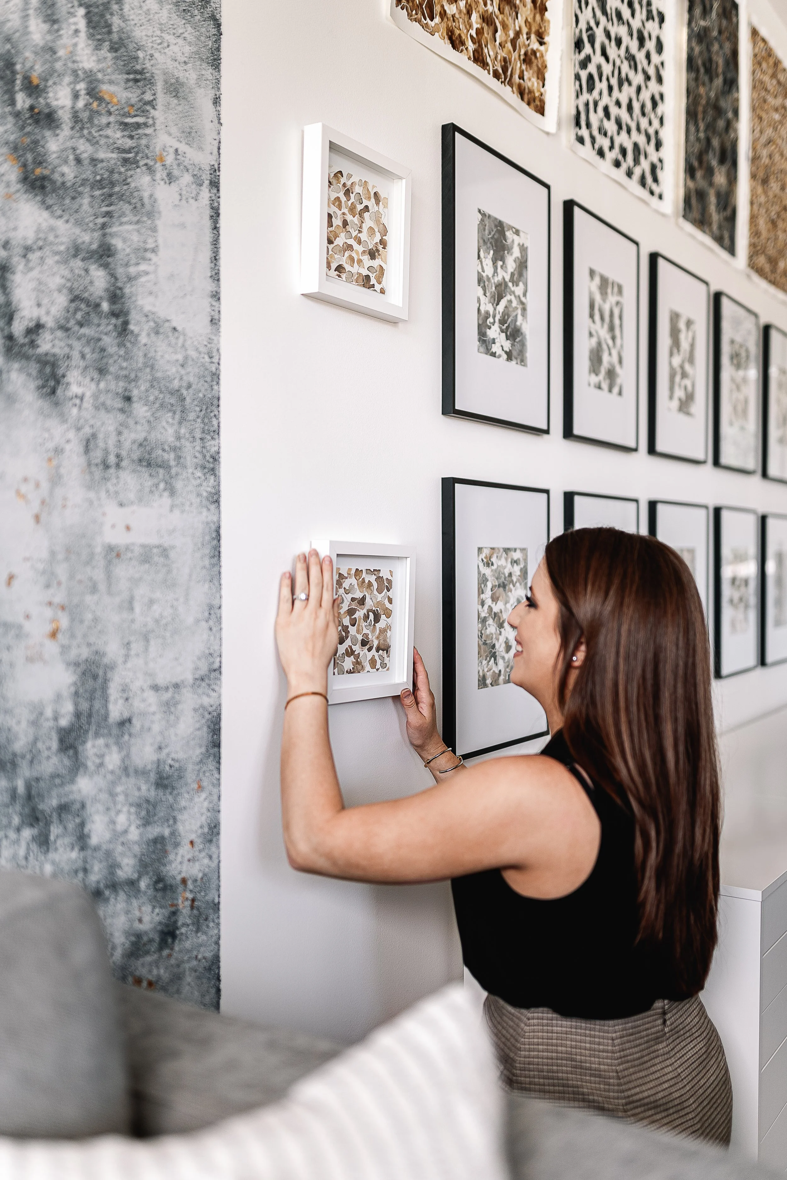 Abstract artist hanging artwork on a gallery wall with multiple framed original paintings in neutral tones