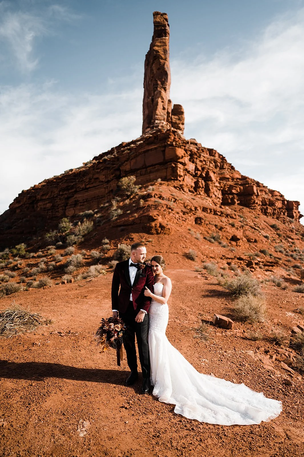 Wedding couple standing on desert terrain with large red rock formation in the background. The bride is in a white wedding gown holding a bouquet, and the groom is in a tuxedo. The sky is partly cloudy.