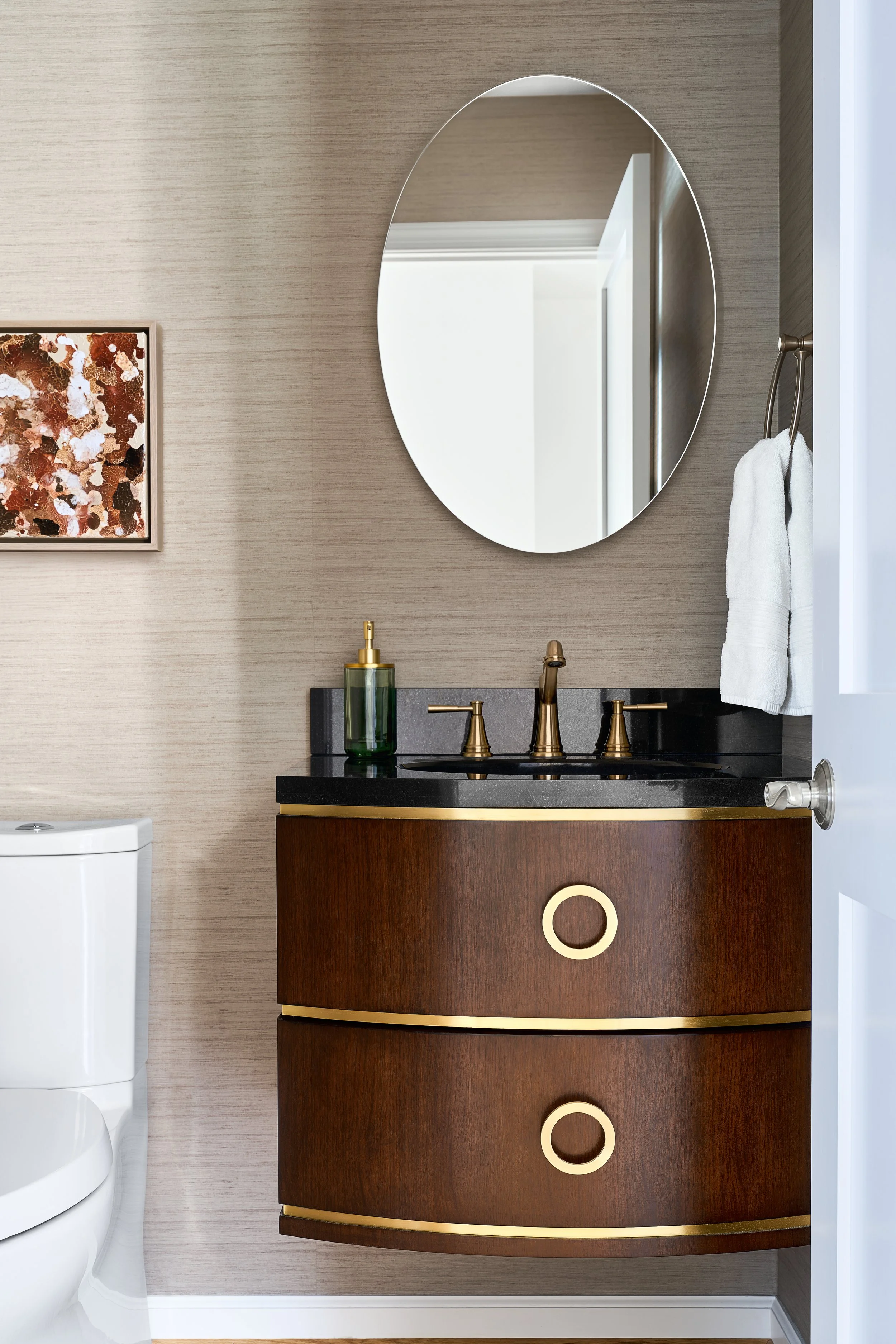 A bathroom with a brown wooden vanity with gold accents, a black countertop, a brass faucet, a round mirror, a white towel, an abstract piece of original wall art on canvas, and a white toilet.