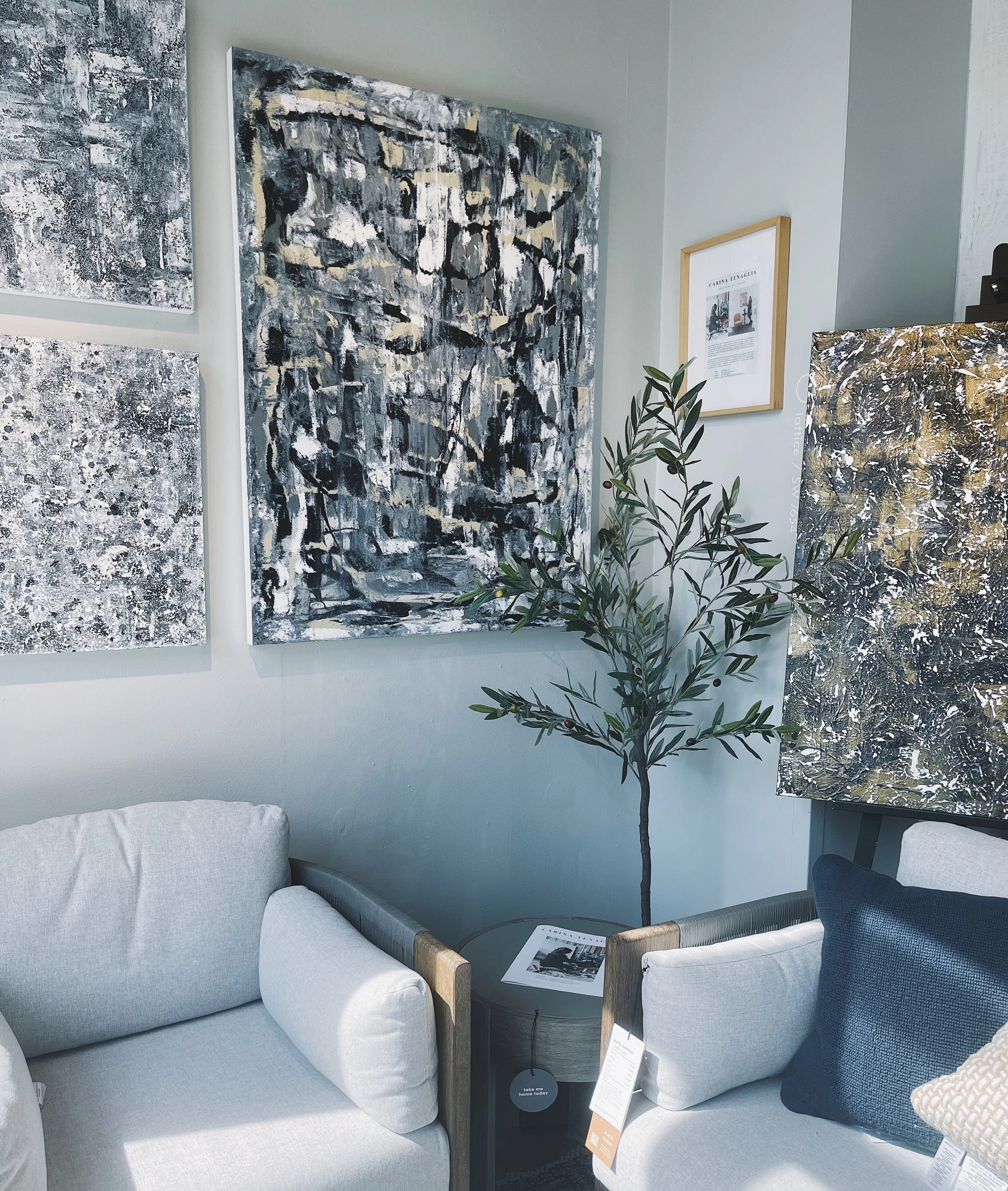 A cozy living room staged at West Elm DC with original abstract artwork on the wall, white sofa with beige and navy cushions, a small indoor plant, and a side table with a magazine.