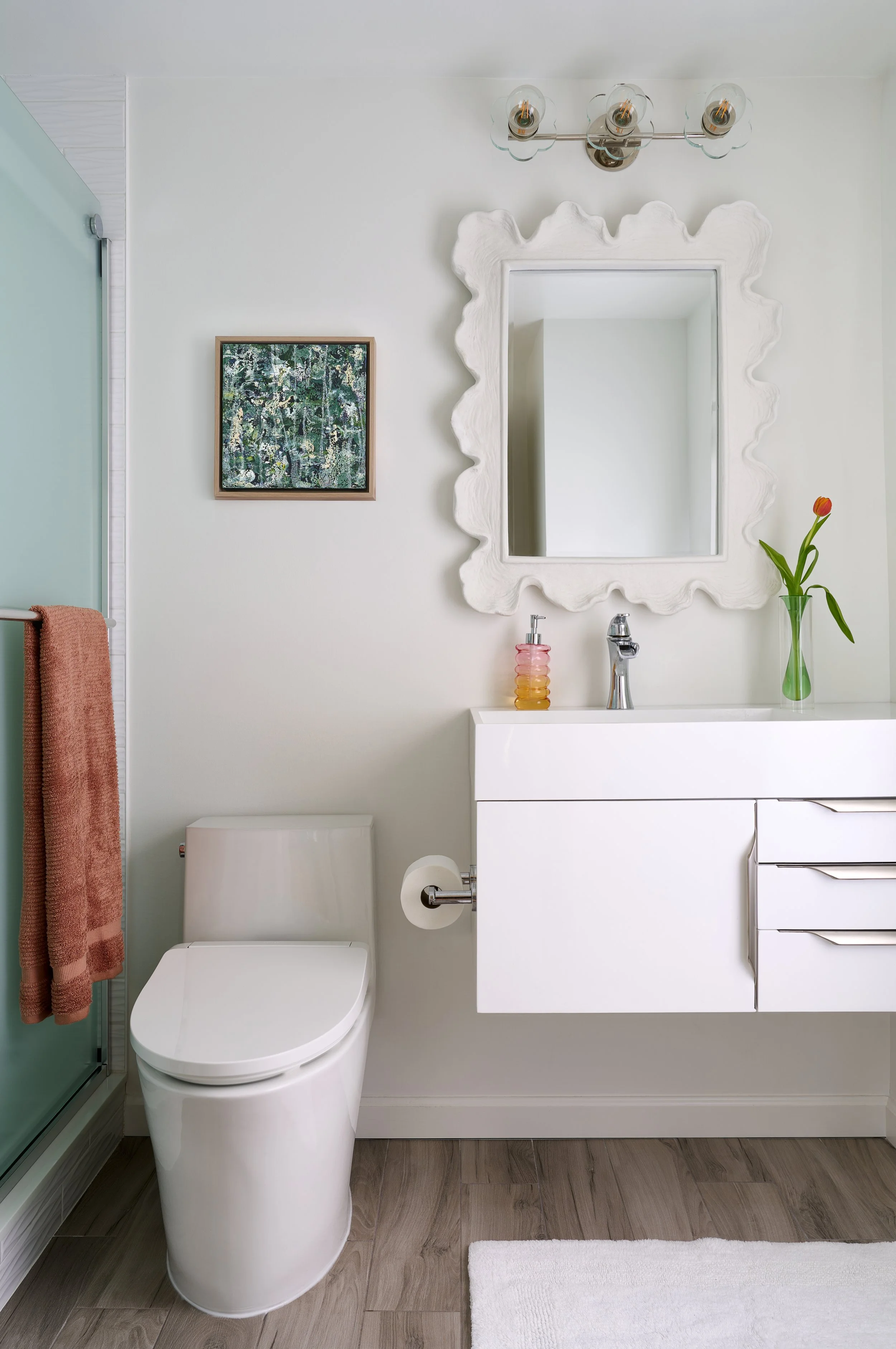 Modern bathroom with white vanity, large mirror, decorative light fixture, textured original framed green artwork, vase with a tulip, and a toilet.