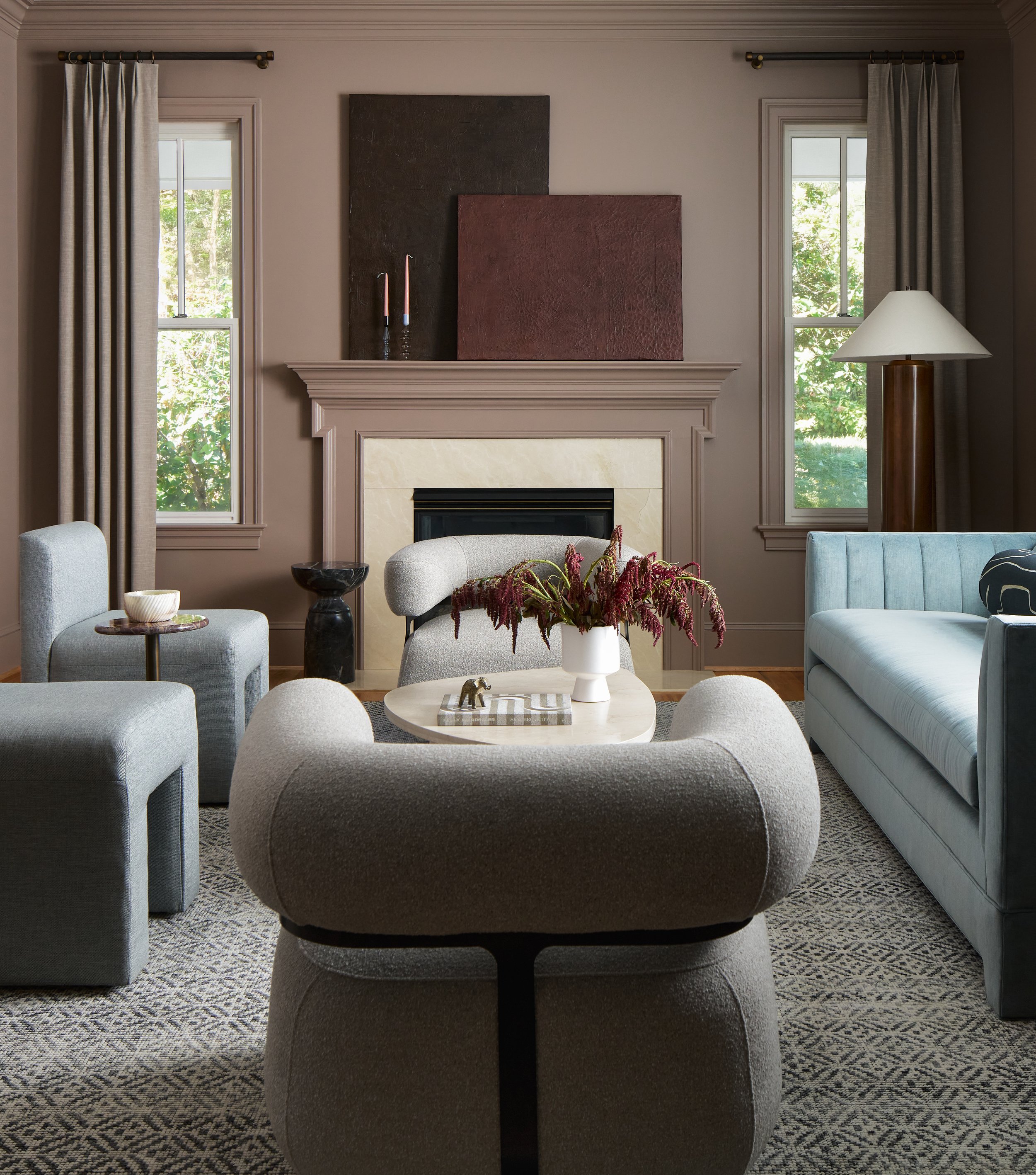 A cozy living room with a fireplace, large windows, and modern furniture. Decorative items include two oversized, moody, warm, textured abstract paintings above the fireplace.