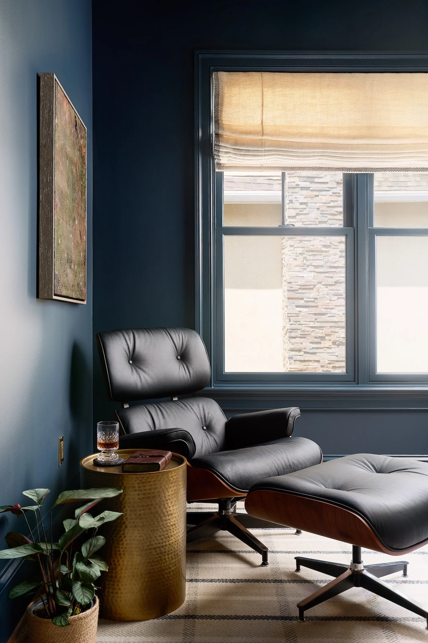 Modern corner of a moody study with a black leather lounge chair and matching ottoman, a gold side table with a glass of drink and a book, a potted plant, a large window with beige shades, and a framed textured original artwork on a dark blue wall.