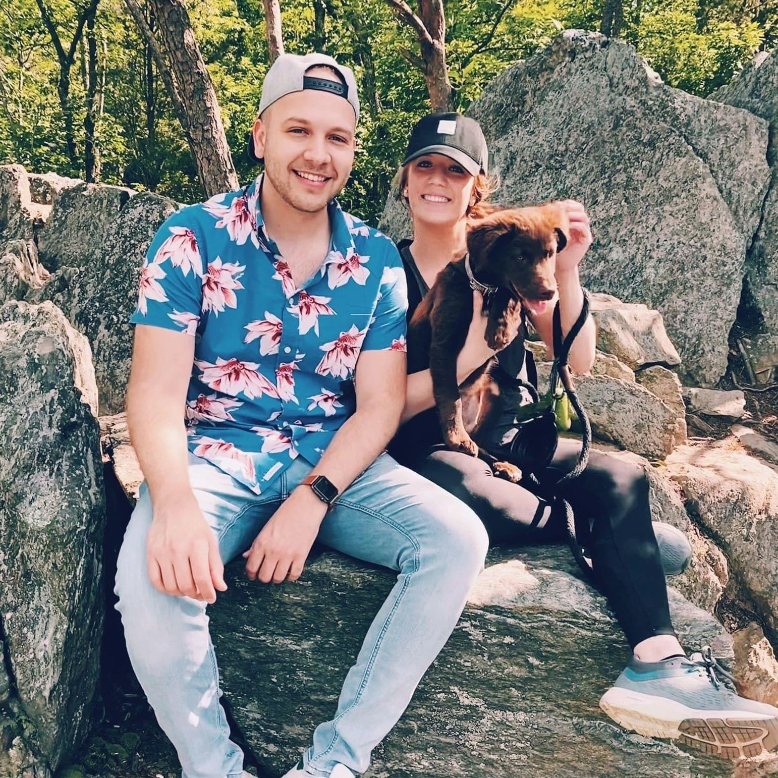 Two people and a dog sitting on rocks in a wooded area, smiling at the camera.
