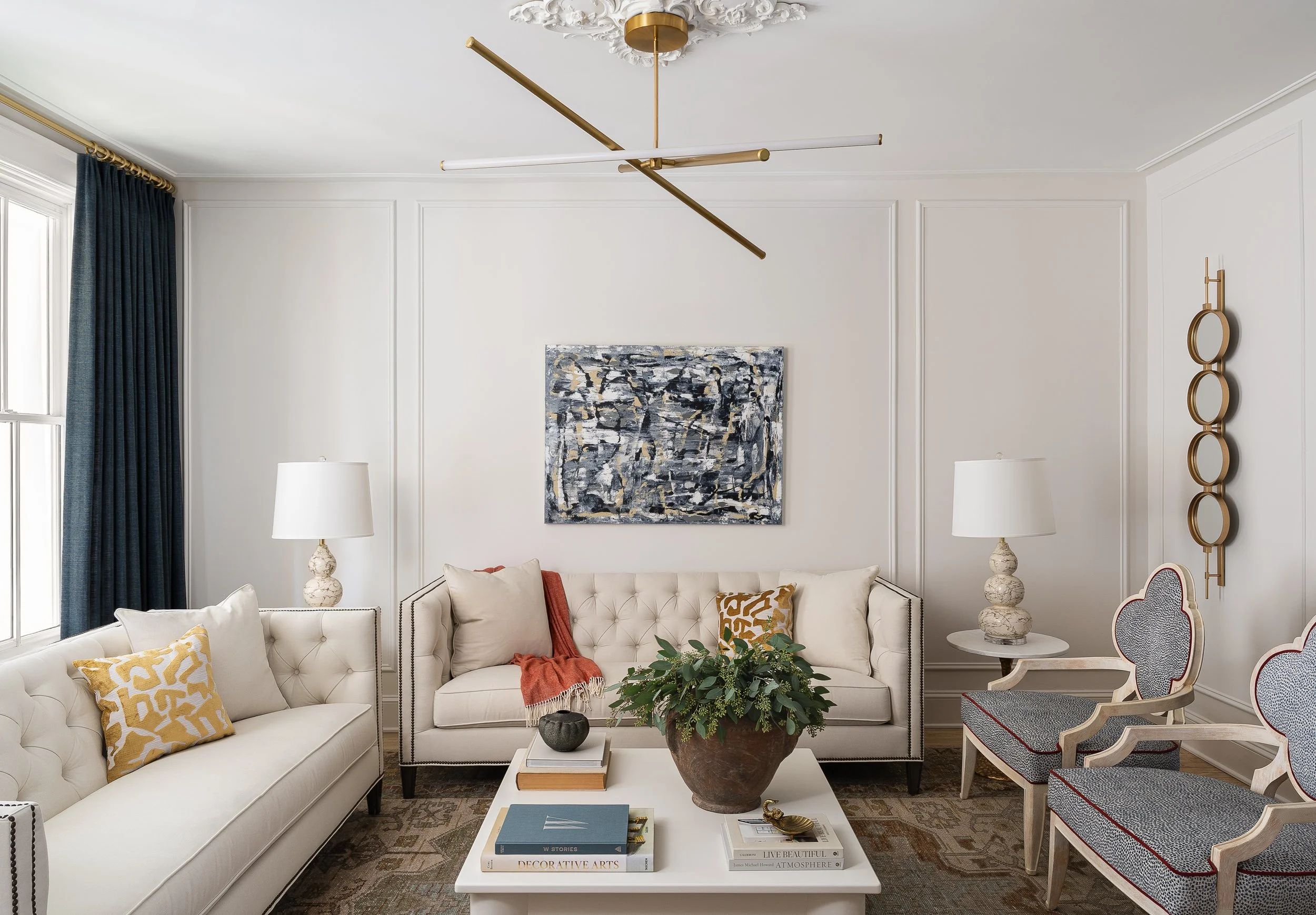 A modern designer living room with white walls, a large window with blue curtains, two white sofas, and two armchairs with patterned fabric. The main wall features an oversized original abstract painting on canvas.