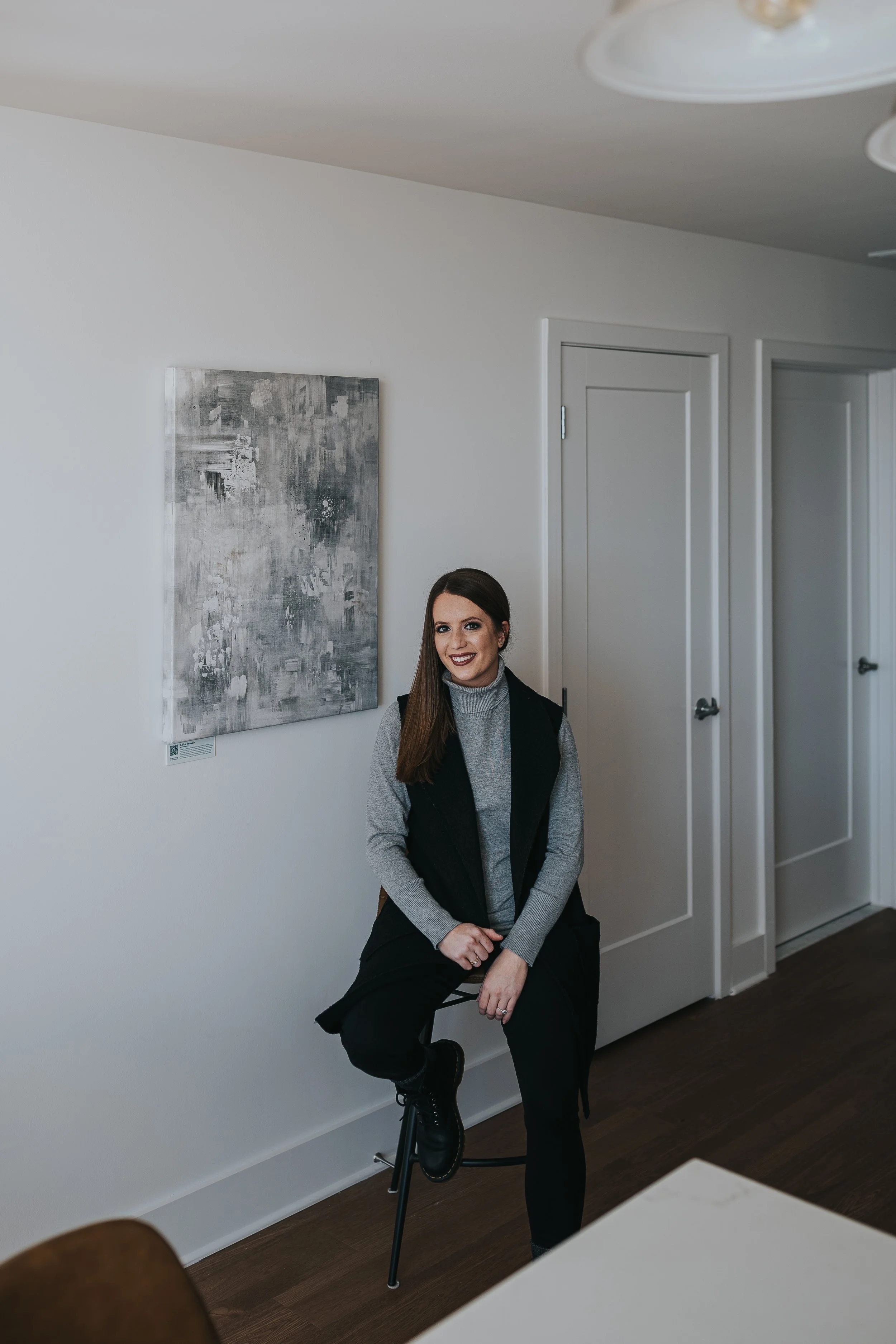 Female DC area artist with long brown hair, wearing a gray turtleneck and black vest, sitting on a stool in a minimalistic room with white walls, dark wooden floor, and abstract art on the wall, licensed to hotel for commercial project.