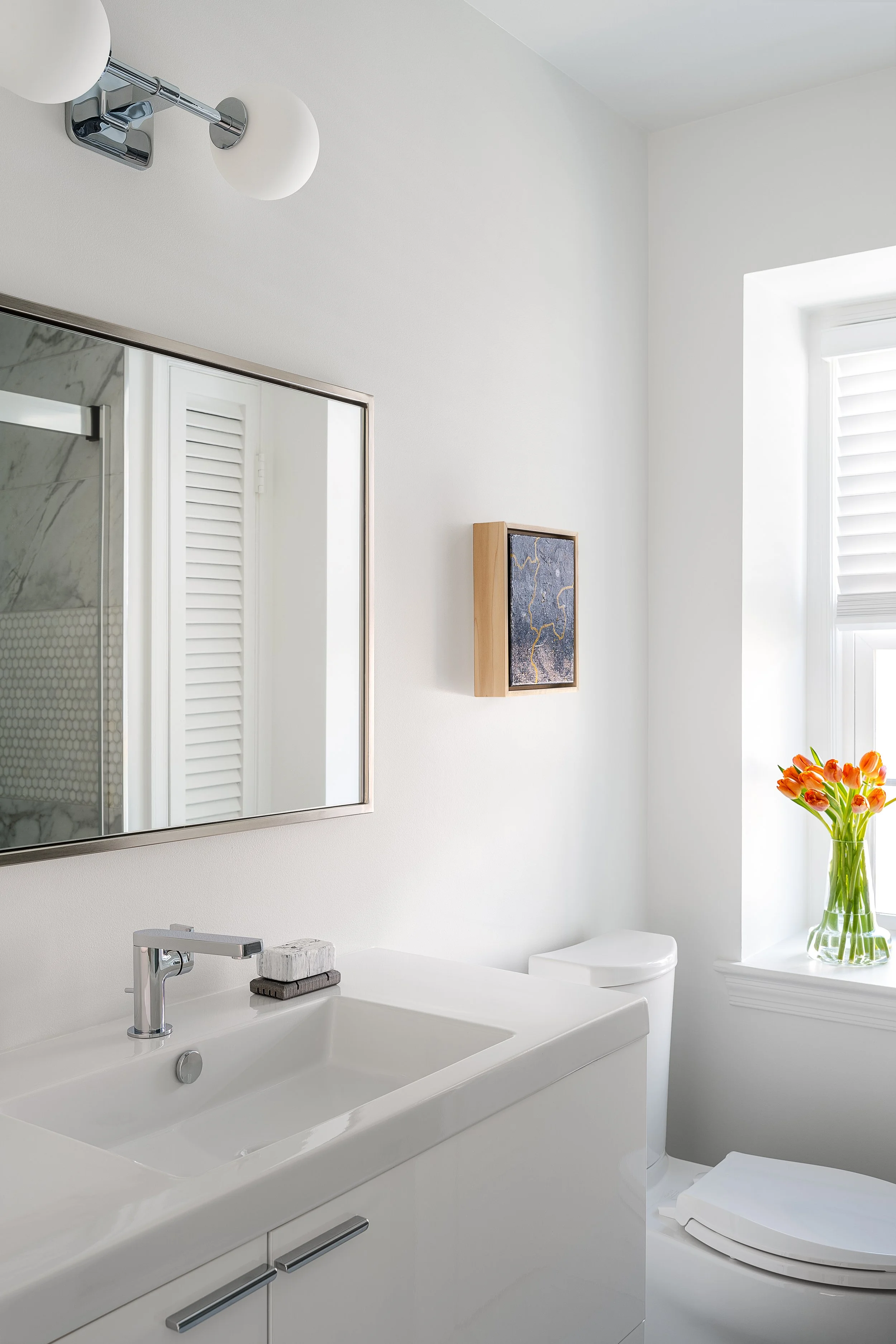 A modern, minimalist bathroom with a white vanity, a rectangular mirror, a wall-mounted light fixture with two round bulbs, a small framed artwork on the wall, a white toilet, and a window with a flower vase containing orange tulips.